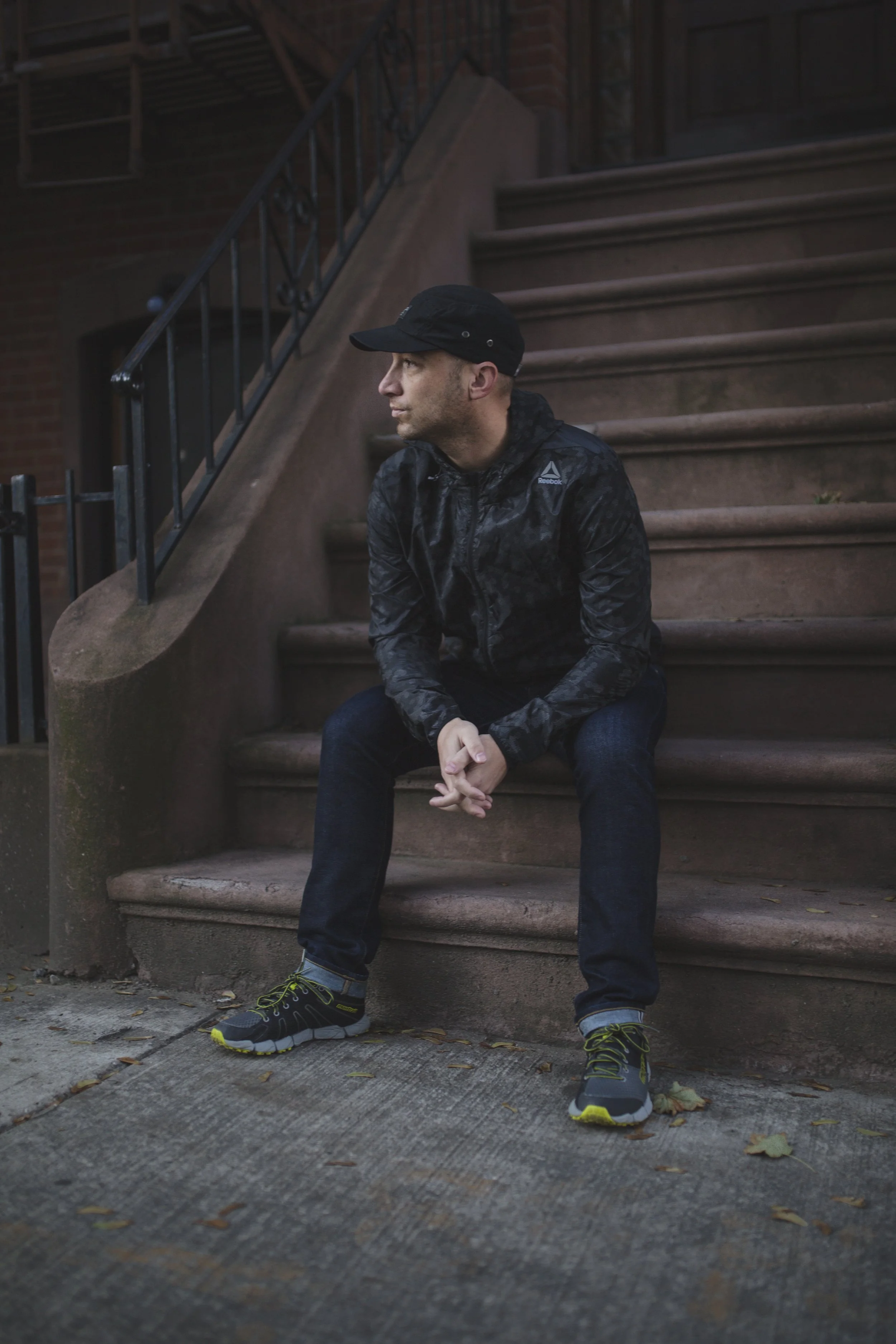 A man sitting on the steps of a brownstone building, wearing a black cap, black jacket with Reebok logo, dark jeans, and running shoes, looking to his left with a thoughtful expression. Reebok.