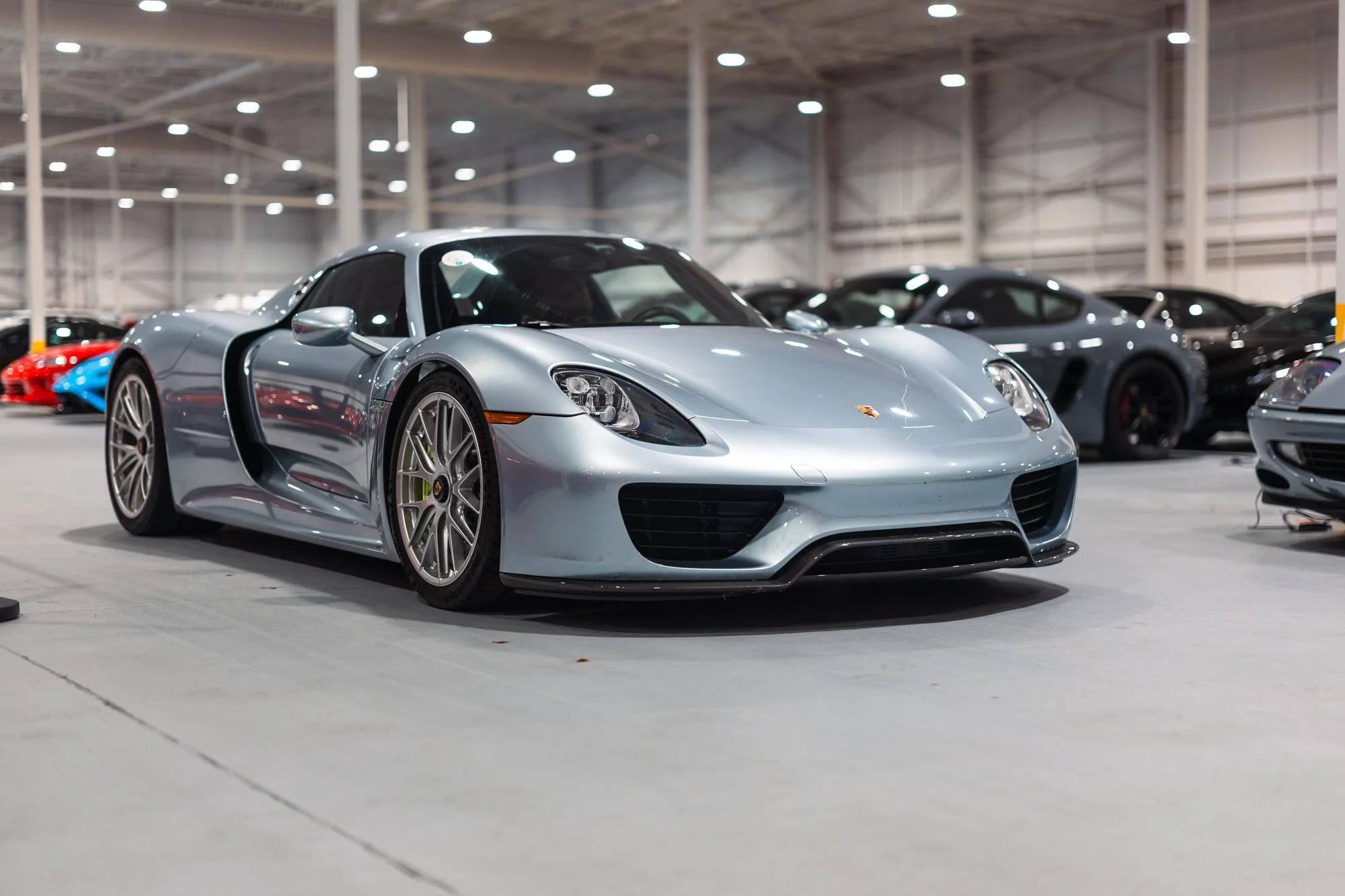 A silver Porsche 918 sports car parked in an indoor showroom with other high-end cars visible in the background.