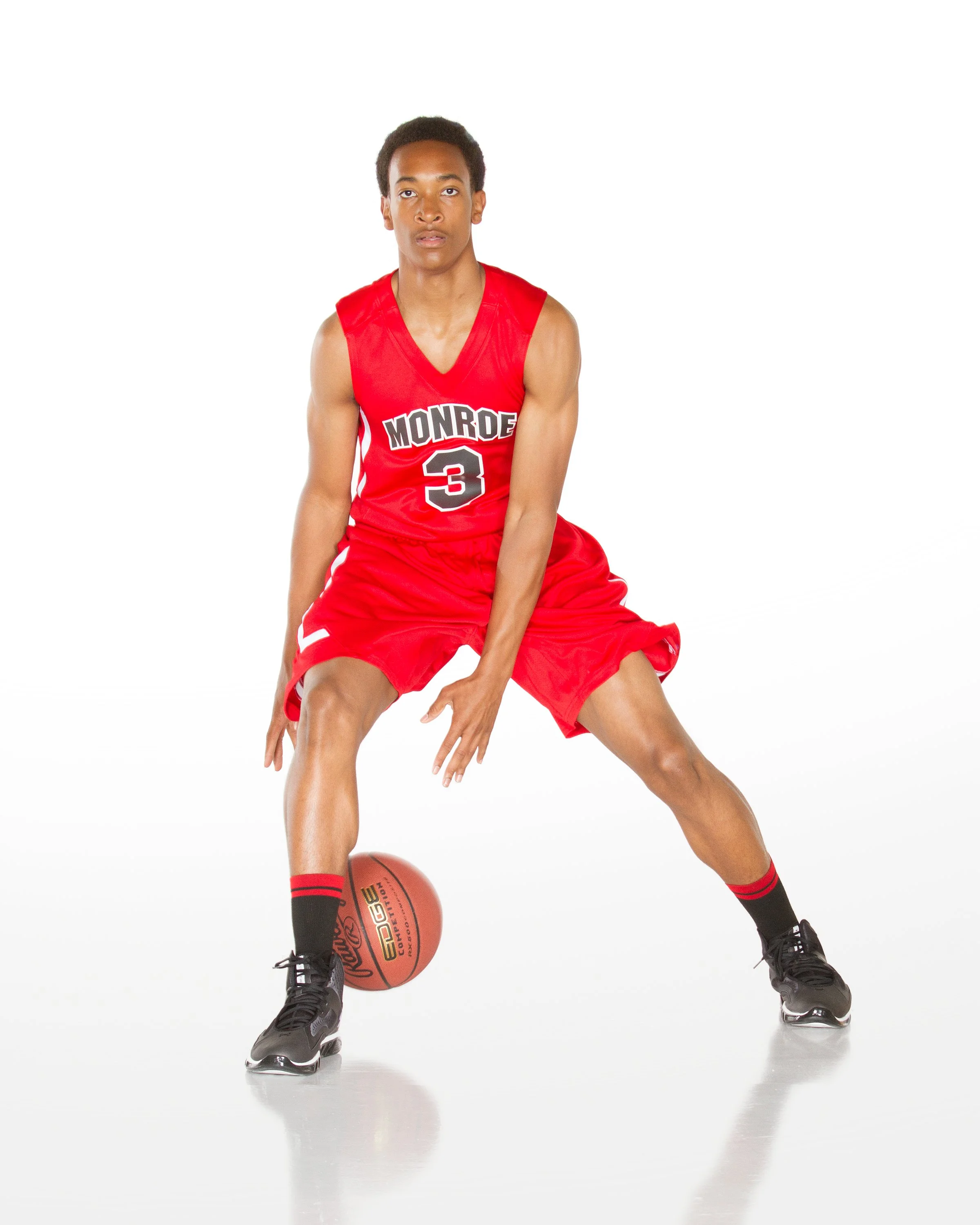 Young male basketball player in red Monroe uniform with number 3, kneeling on ice rink with one hand touching the ground, holding a basketball with the other, wearing black ice skates.