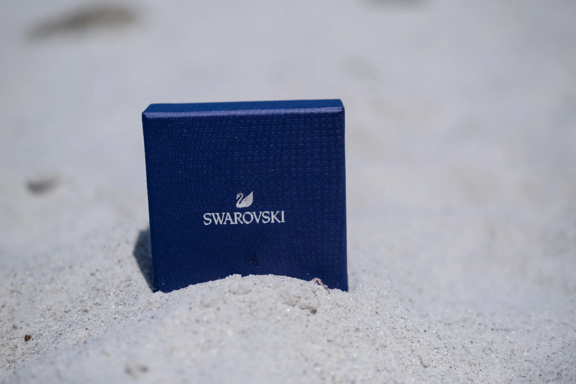 Blue Swarovski jewelry box placed on sandy beach.