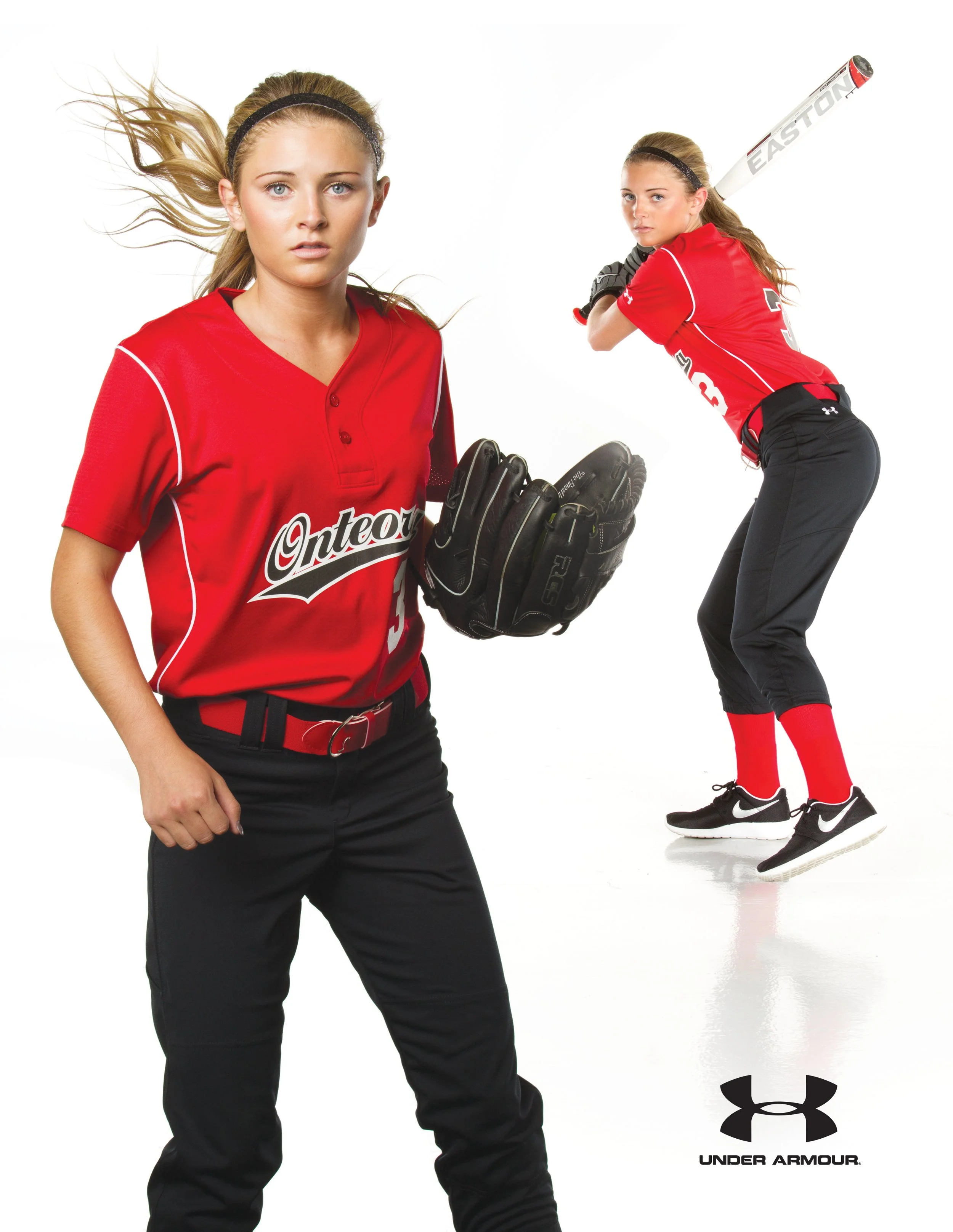 Two young female softball players wearing red jerseys, black pants, and black gloves against a white background. One is holding a softball glove, and the other is in a batting stance with a bat over her shoulder. The Under Armour logo is visible at t