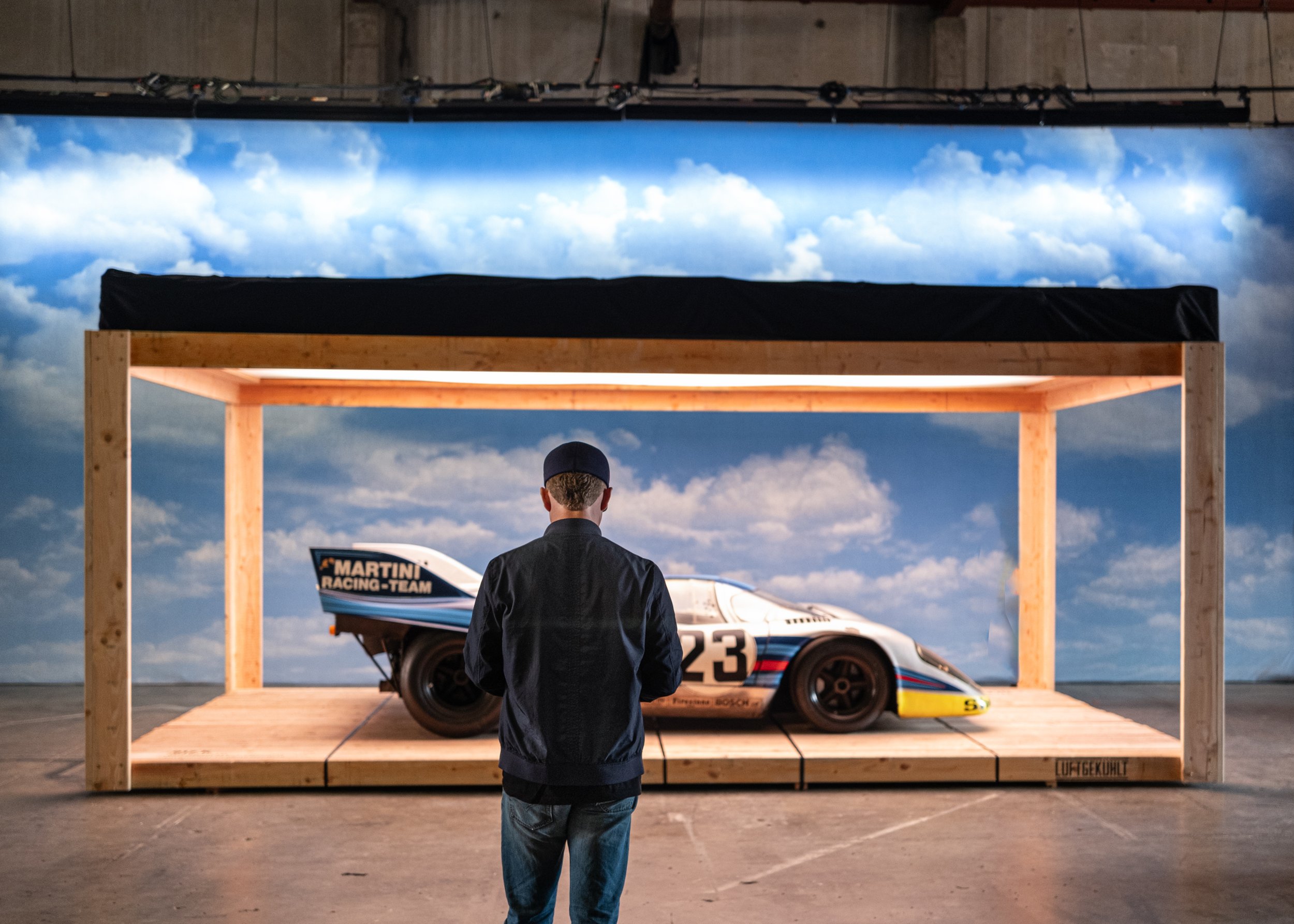 A person is standing inside a wooden frame on a platform, facing away from the camera, with a race car in front of them. The background shows a sky with clouds.