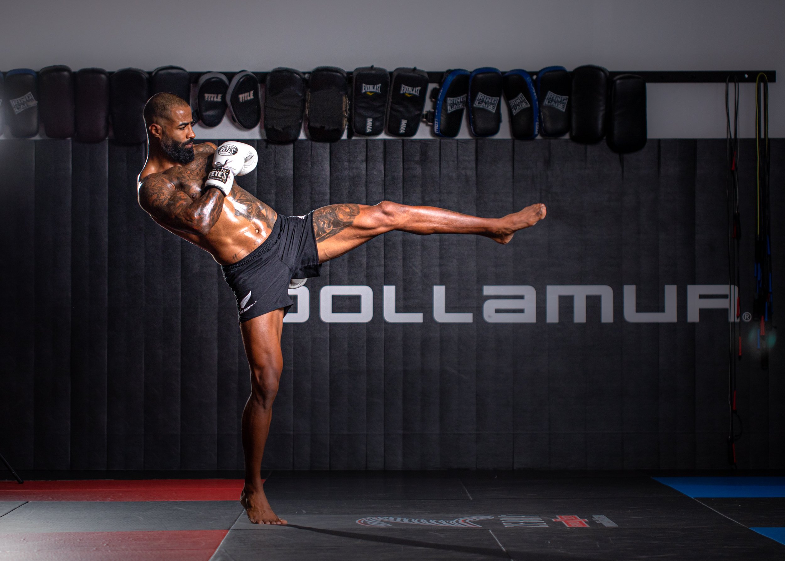 A male martial artist practicing a high kick in a gym with black padded walls and stored boxing gloves hanging on the wall. Cosmo Alexandre, muay thai