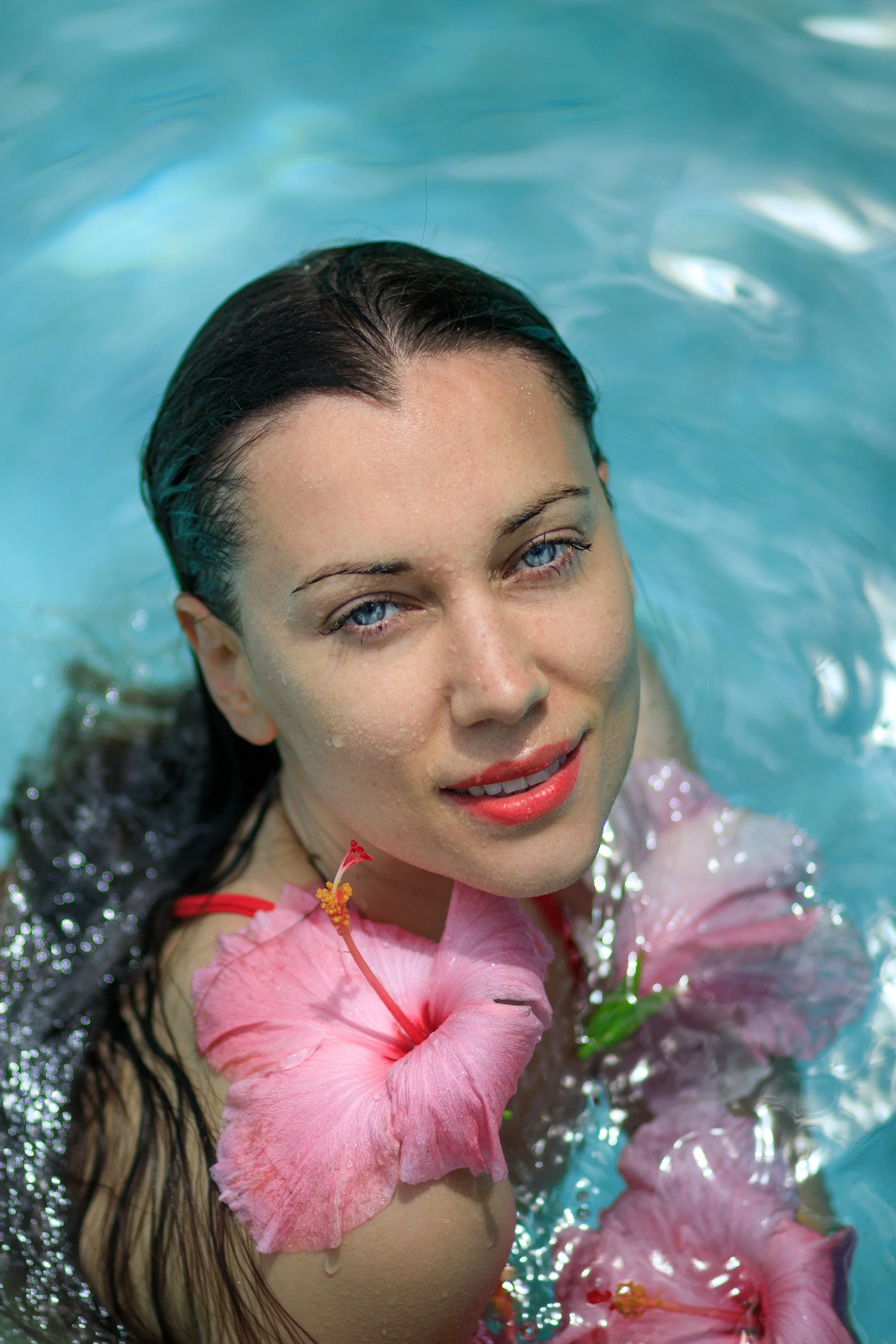 Close-up of a woman with wet dark hair and blue eyes submerged in water, surrounded by pink hibiscus flowers. Vogue italia.