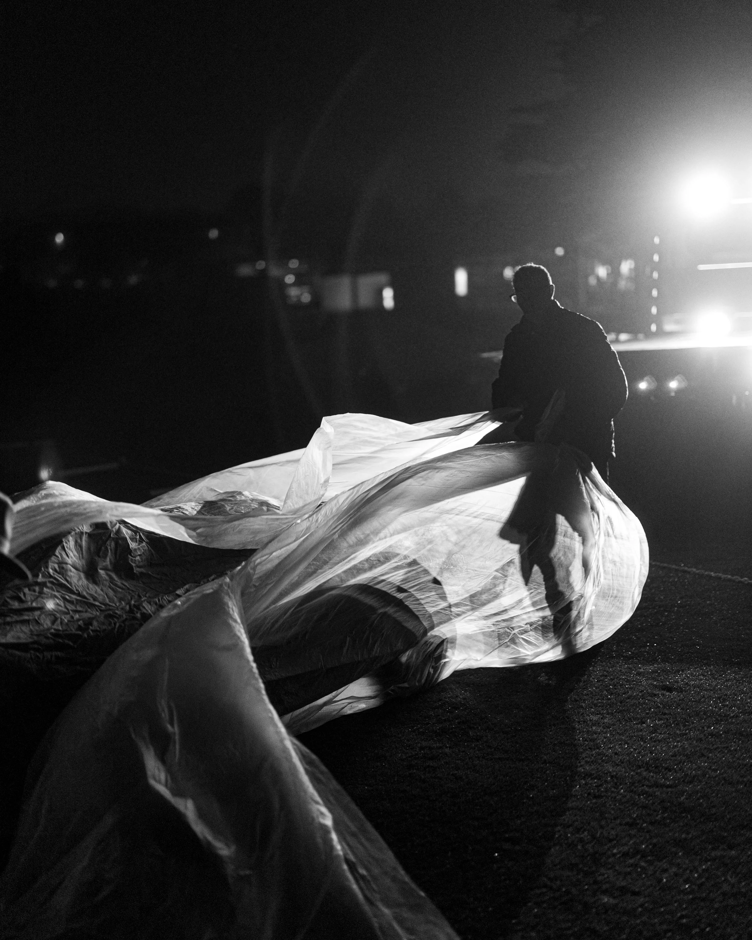 A person working outdoors at night, handling a large plastic sheet or tarp, with bright lights in the background and some blurred vehicles or objects.