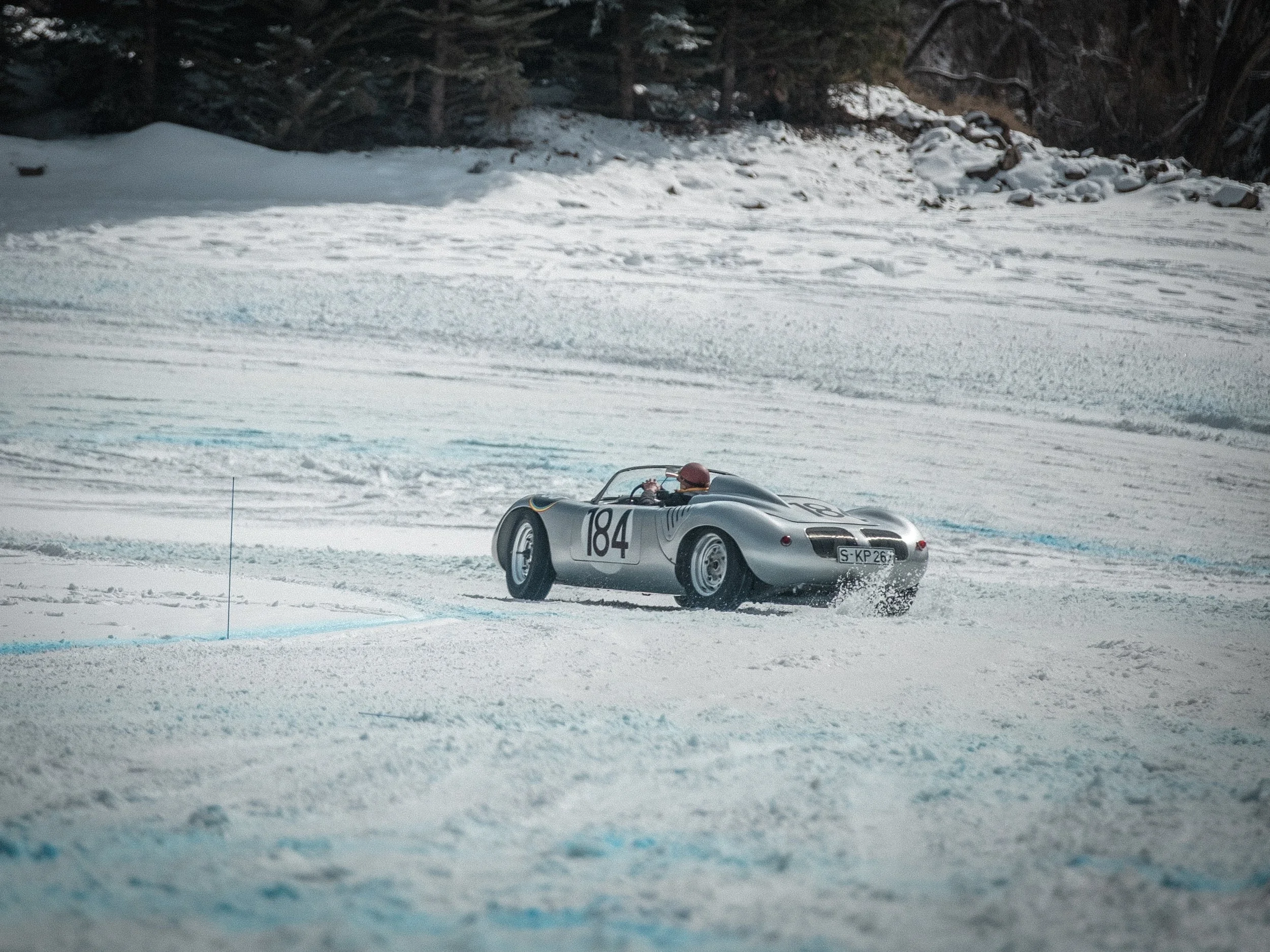 A sleek, vintage silver race car with the number 184 on its side is driving on a snowy track, with snow-covered ground and coniferous trees in the background. Porsche race car, Jeff Zwart driver, Fat Ice Race, Aspen Colorado