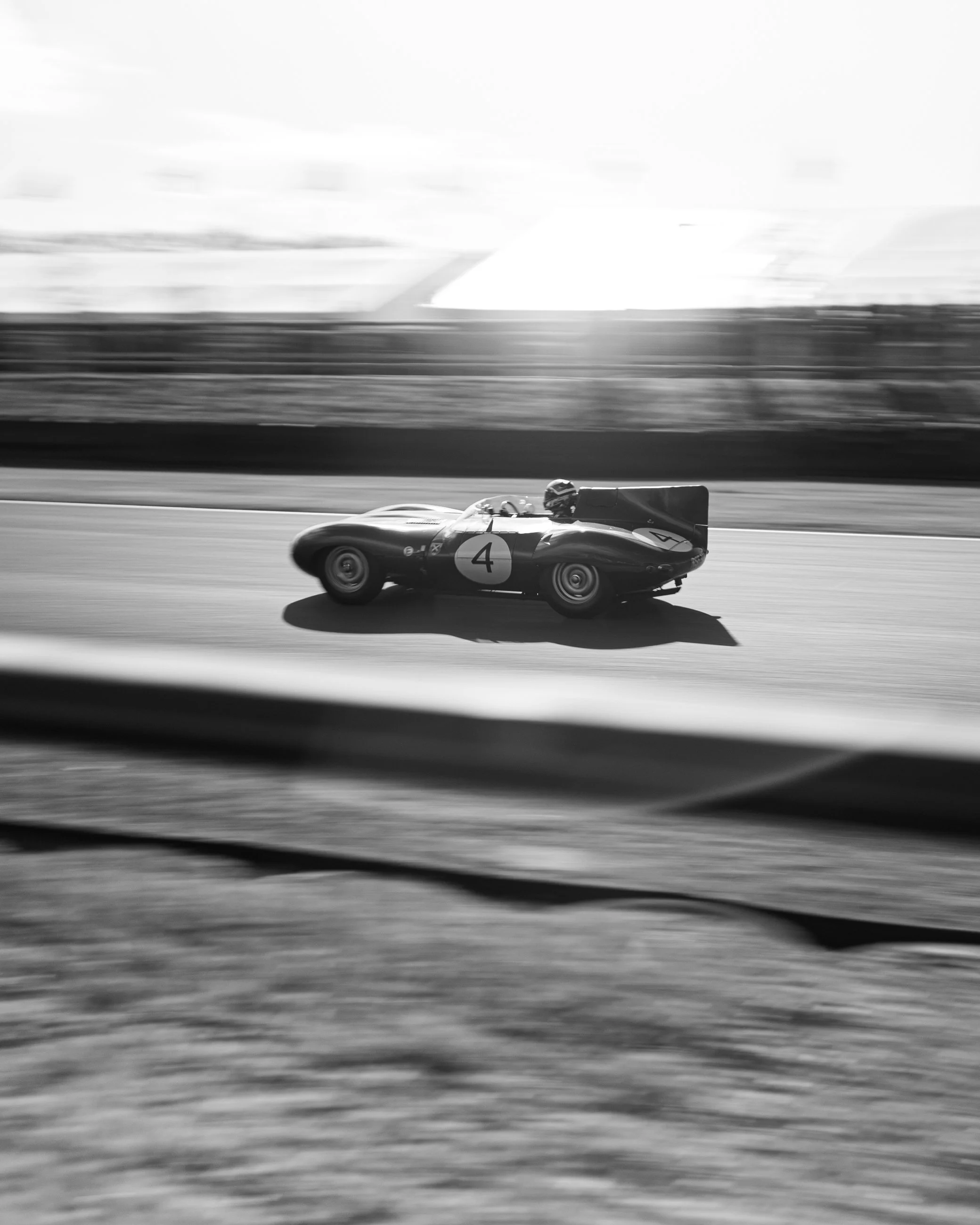 Black and white photo of a race car with the number 4 on its side speeding on a track, with blurred background indicating motion. Goodwood Revival.