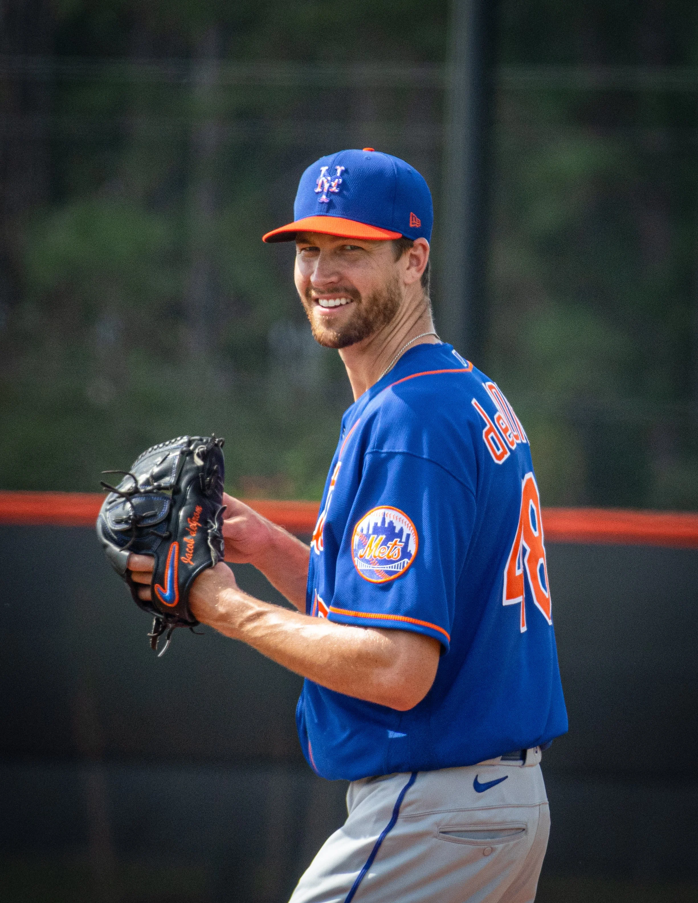 A baseball player wearing a blue NY Mets cap, a blue jersey with orange accents, and white pants with blue piping, standing outdoors on a baseball field. Jacob Degrom