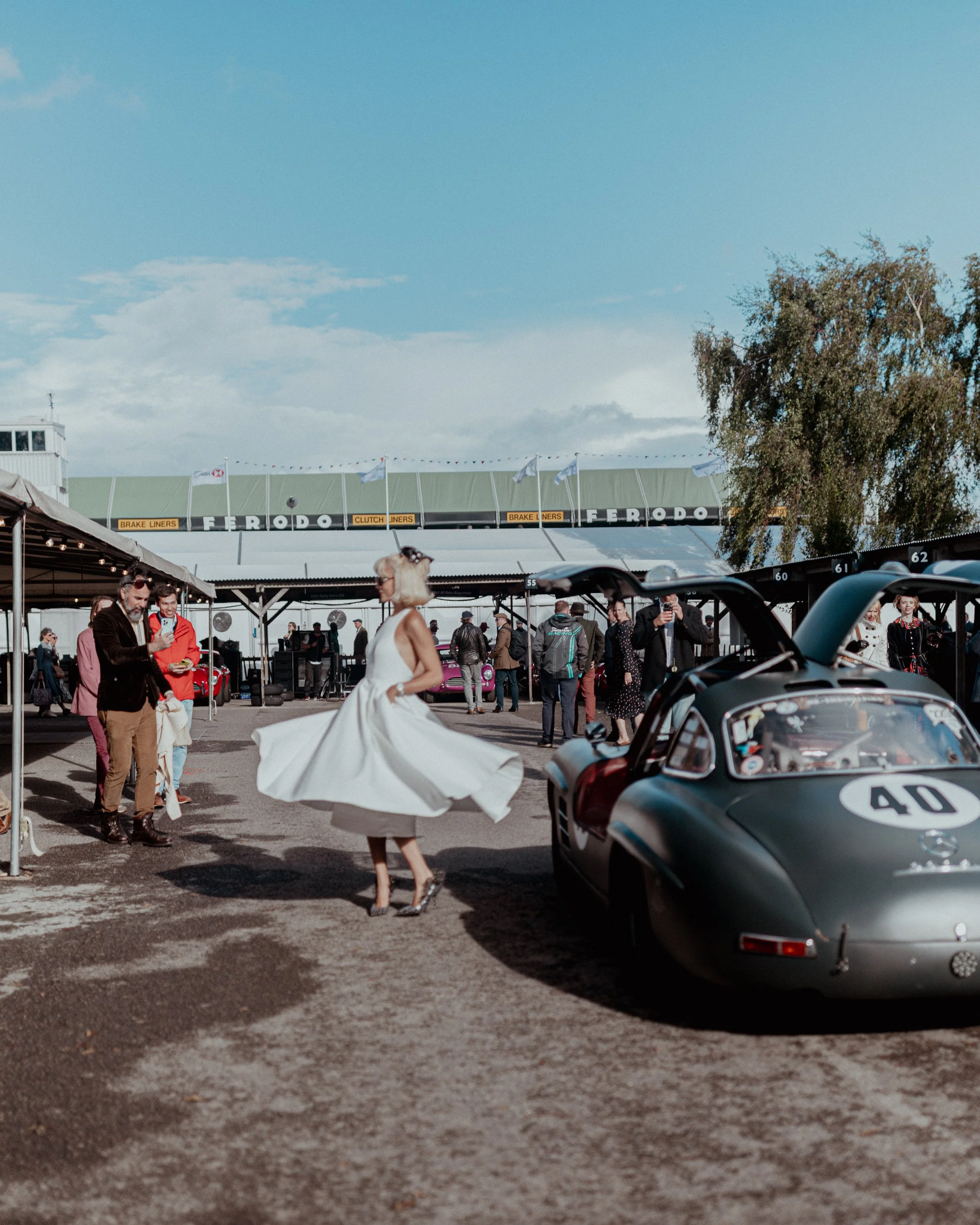 Vintage race car with the number 40 parked near a crowd at a racing event. People are gathered around, some taking photos. A woman in a white dress is walking past, and there are tents and a building structure in the background beneath a partly cloud