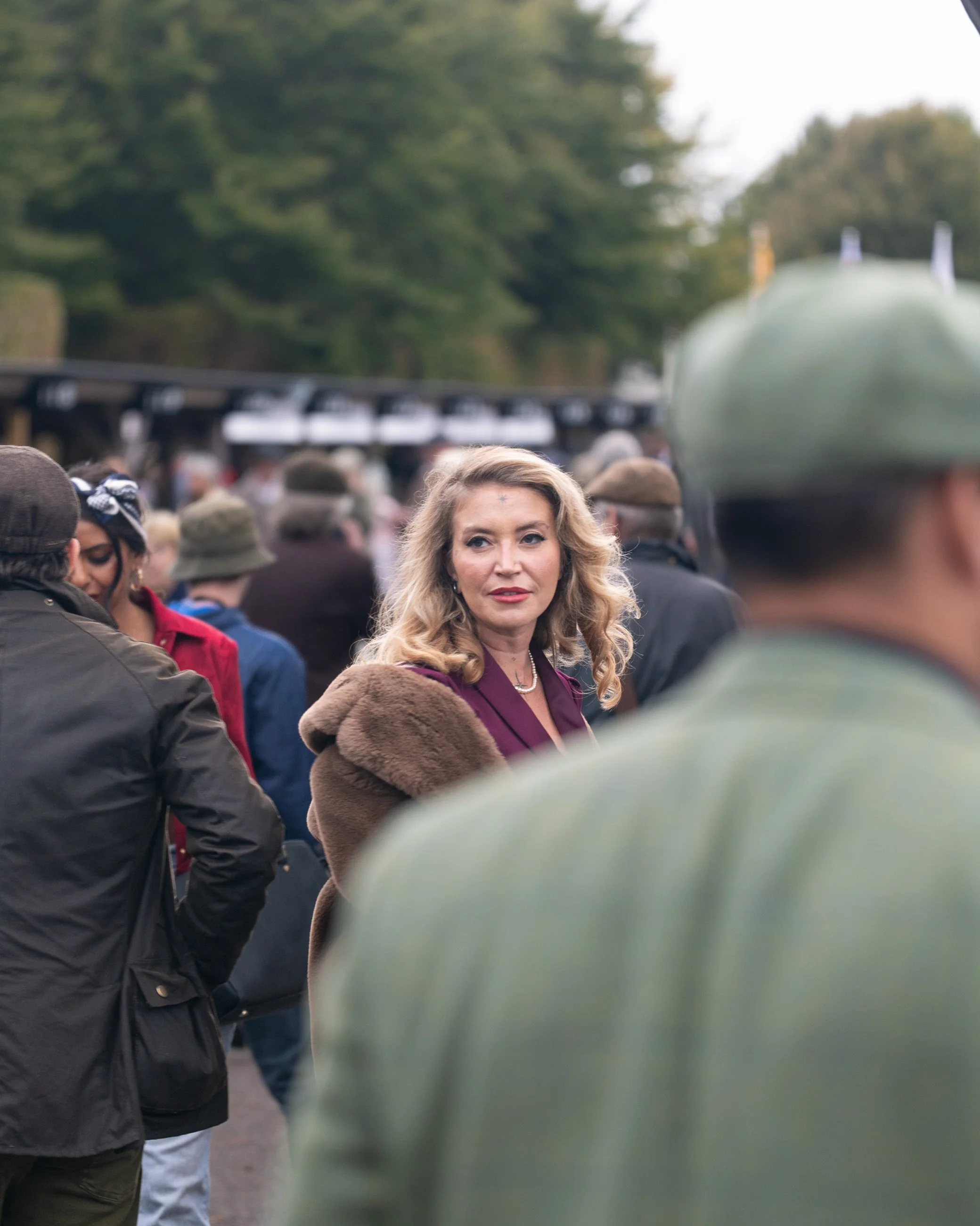 A woman with blonde, wavy hair wearing a burgundy blazer and pearl necklace in a crowd at an outdoor event, with a hazy green background and a person with a green cap in the foreground. Goodwood Revival.