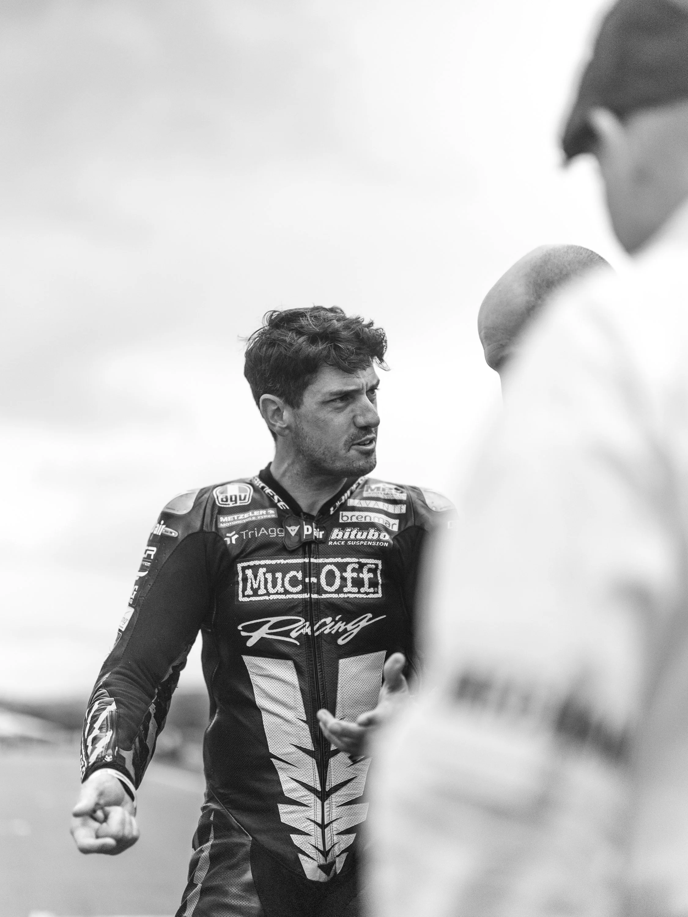 A male race car driver in a racing suit engaging with someone, with a blurred person in the foreground. The race car driver has dark, curly hair and is outdoors against a cloudy sky.