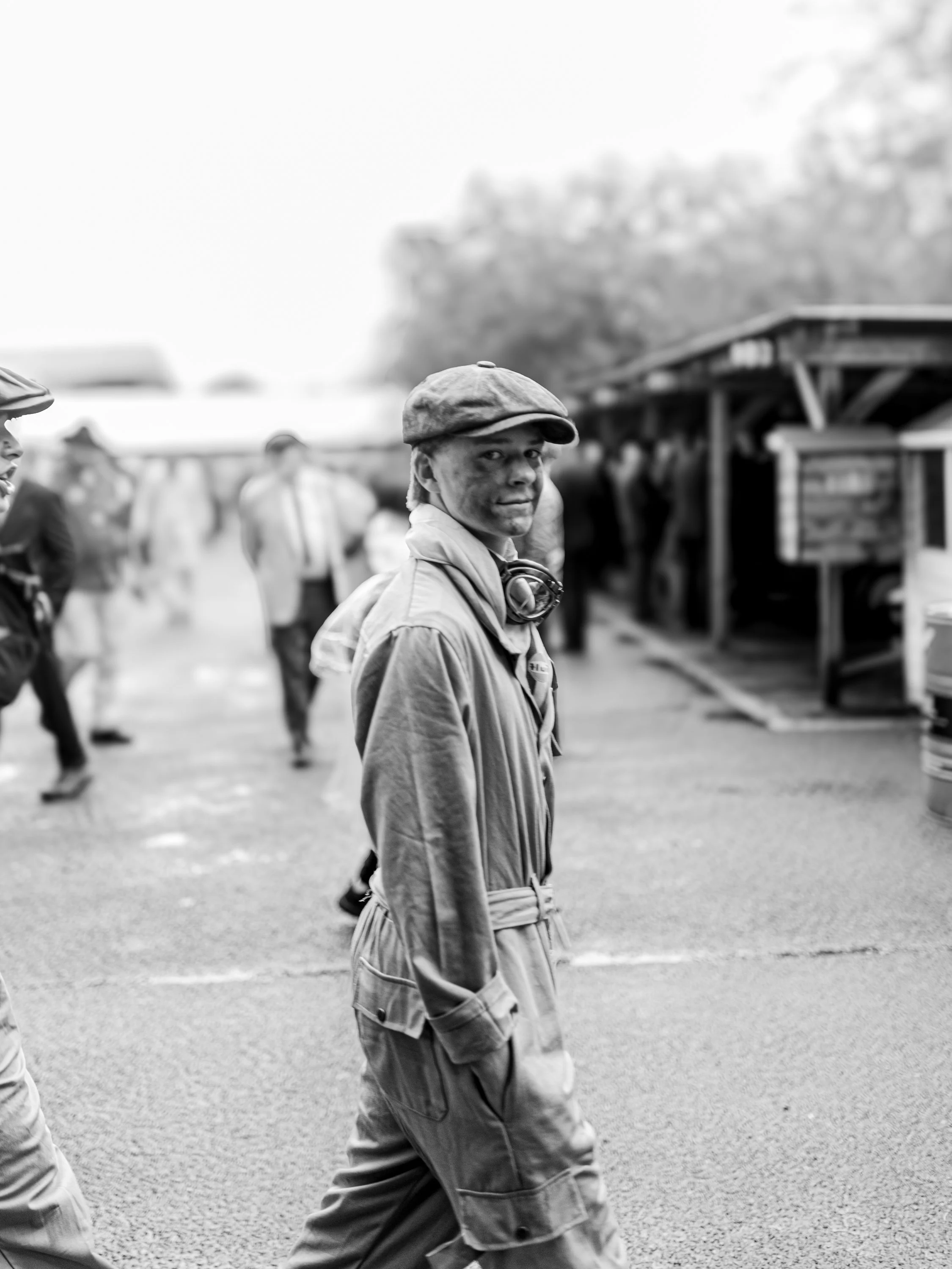 A young man with a hat and headphones around his neck walking outdoors, smiling, with several people and a wooden structure in the background.