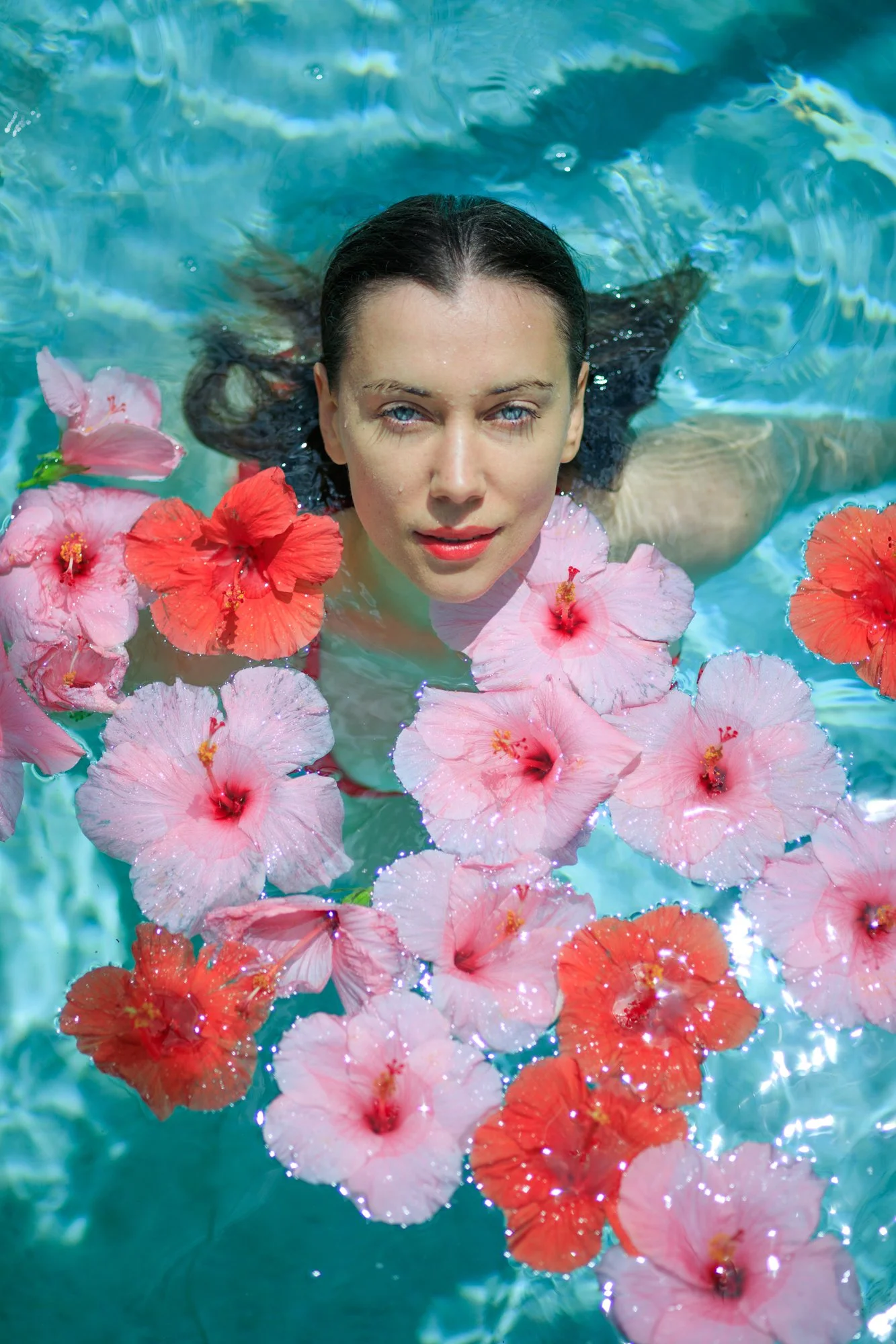 A woman with wet dark hair and blue eyes floating in a pool with pink and red hibiscus flowers surrounding her. Vogue italia.
