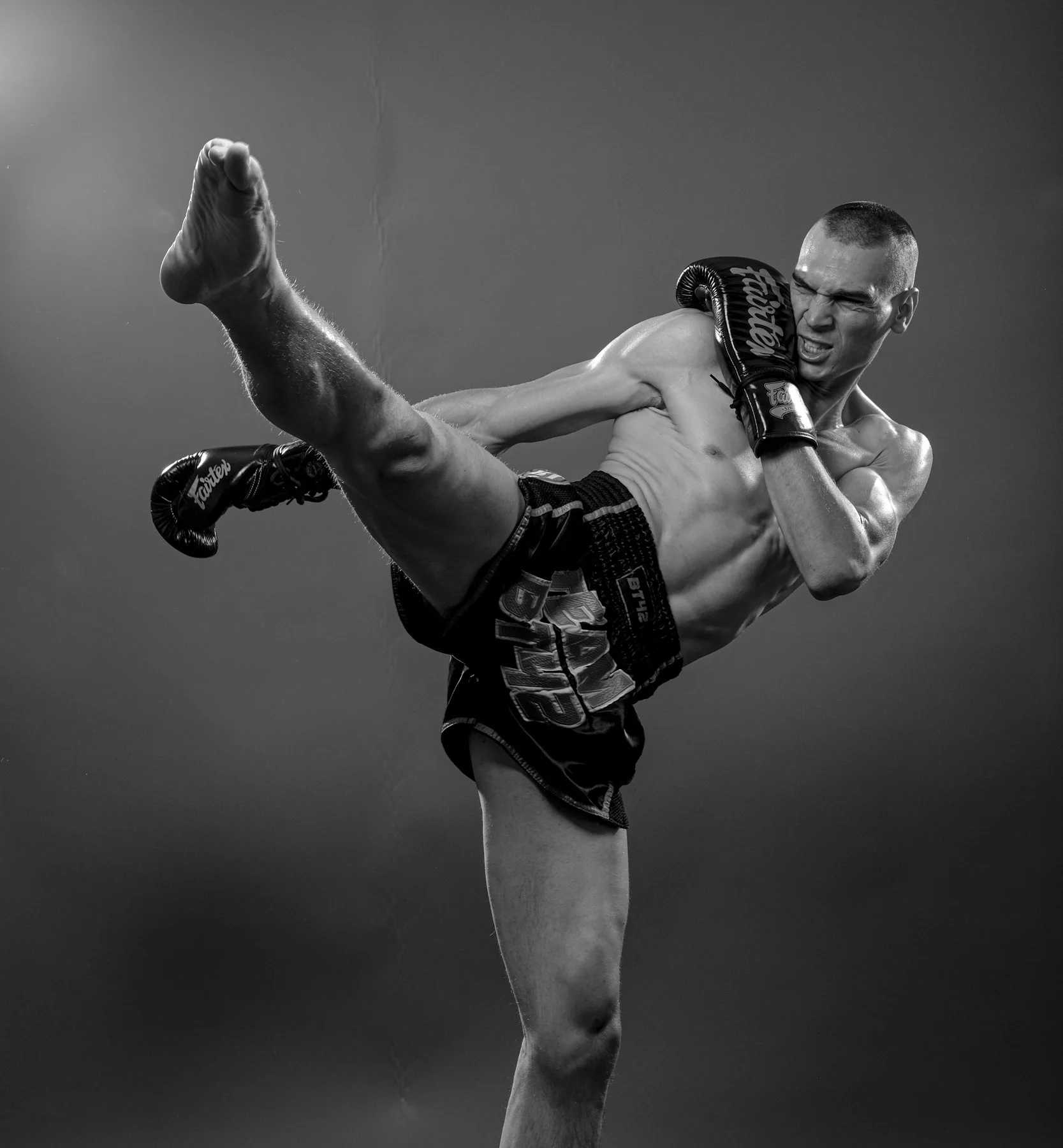Black and white photo of a mixed martial arts fighter delivering a high kick, wearing gloves and athletic shorts.