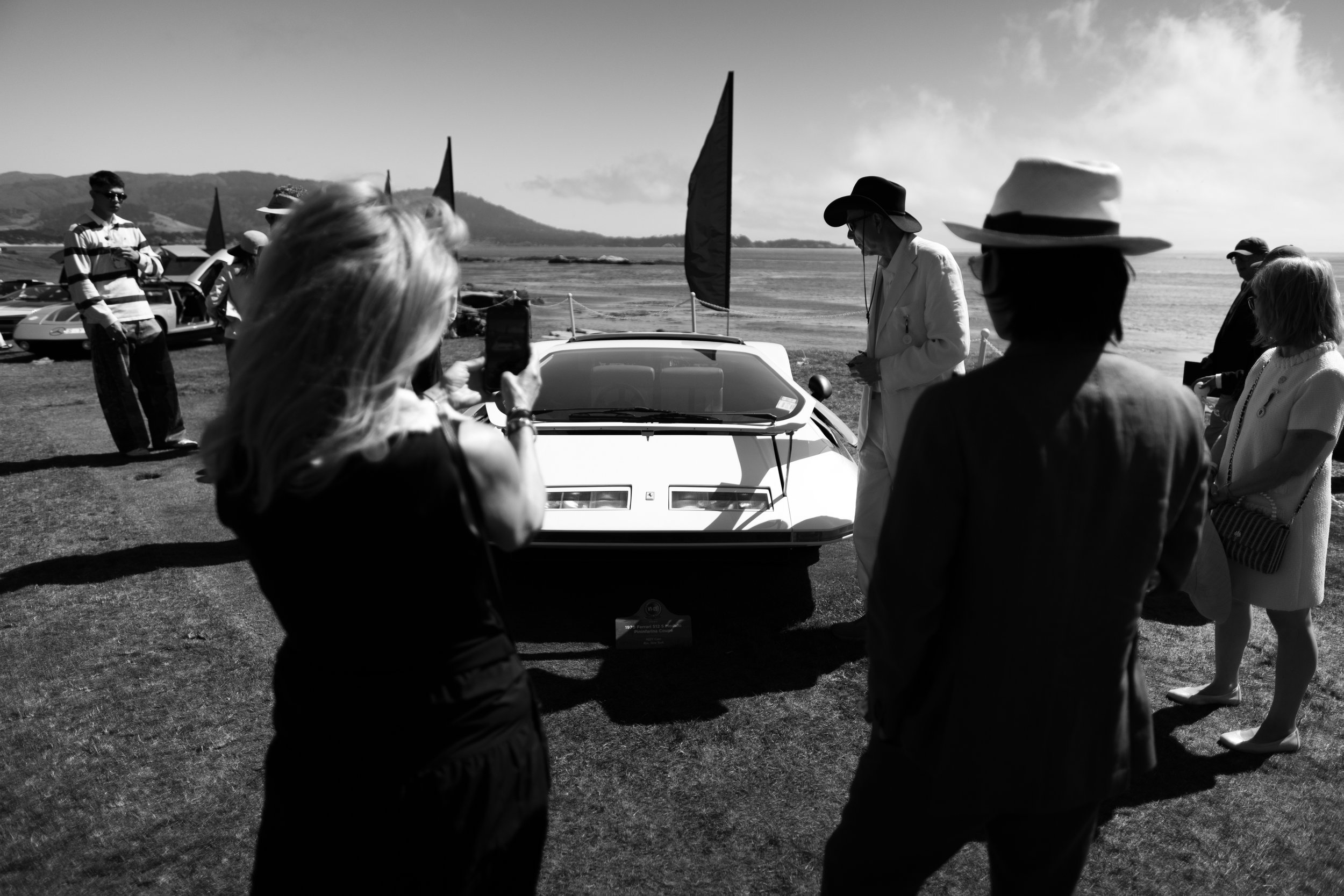 Group of people gathered outdoors around a vintage car, with some individuals taking photos and others chatting, near a scenic body of water and hills.