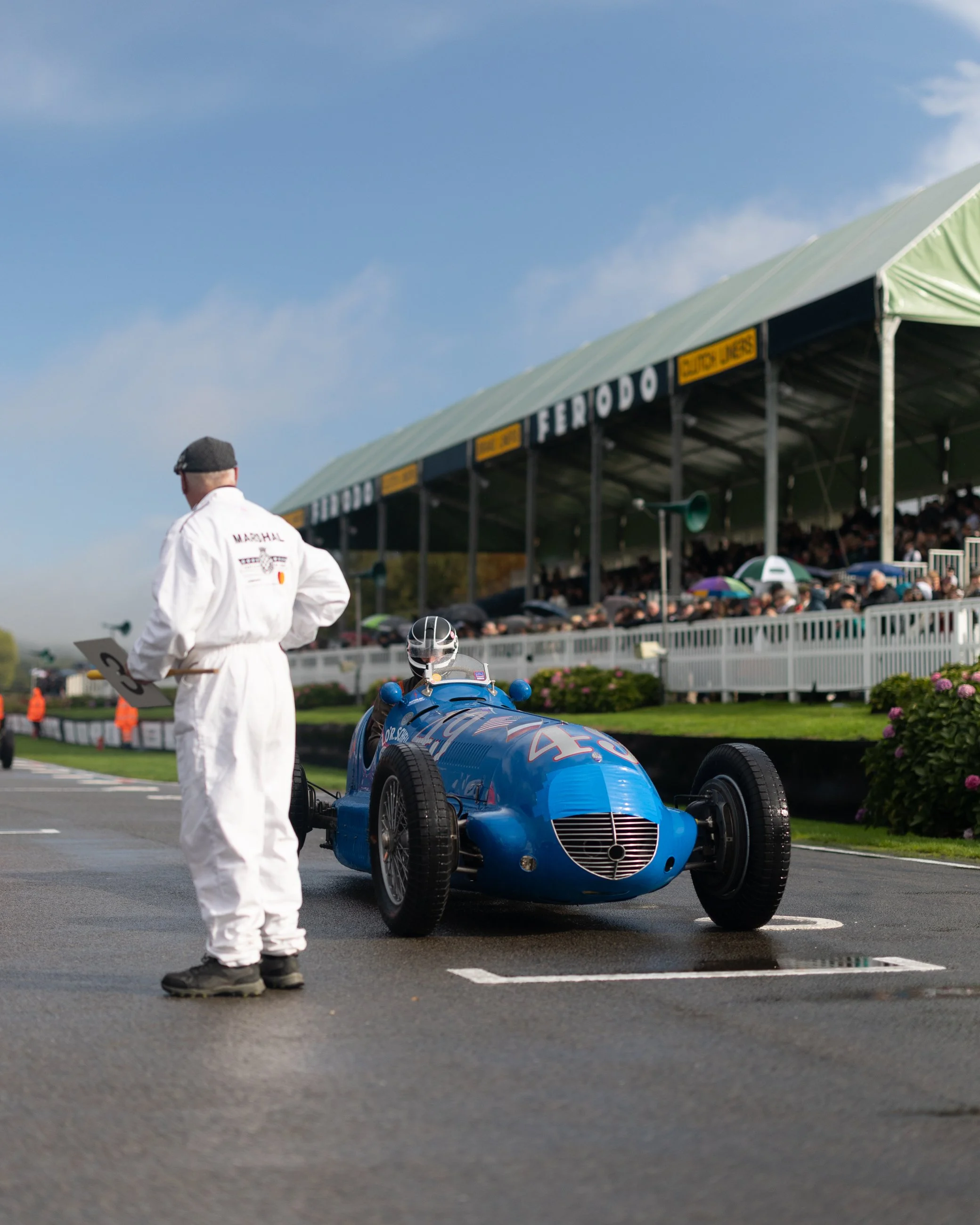 A vintage blue race car on a wet track with a man in white coveralls and a cap standing nearby, with a large spectator stand full of people under an overcast sky.