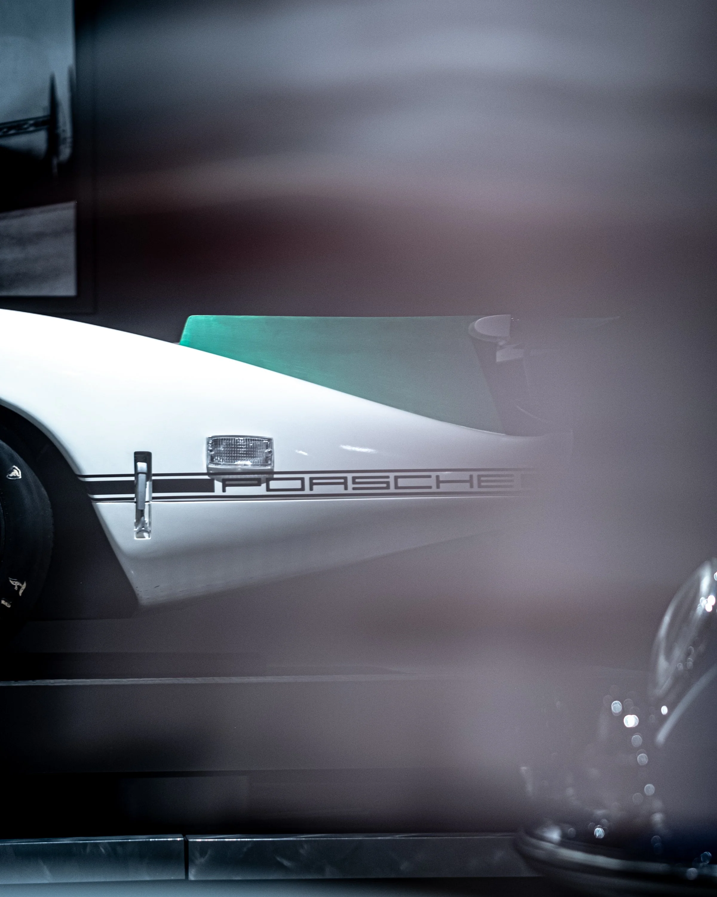Close-up of a white Porsche car, showing part of the front headlight, a black bumper with the Porsche logo, and a chrome side mirror. The background is dark and blurred.