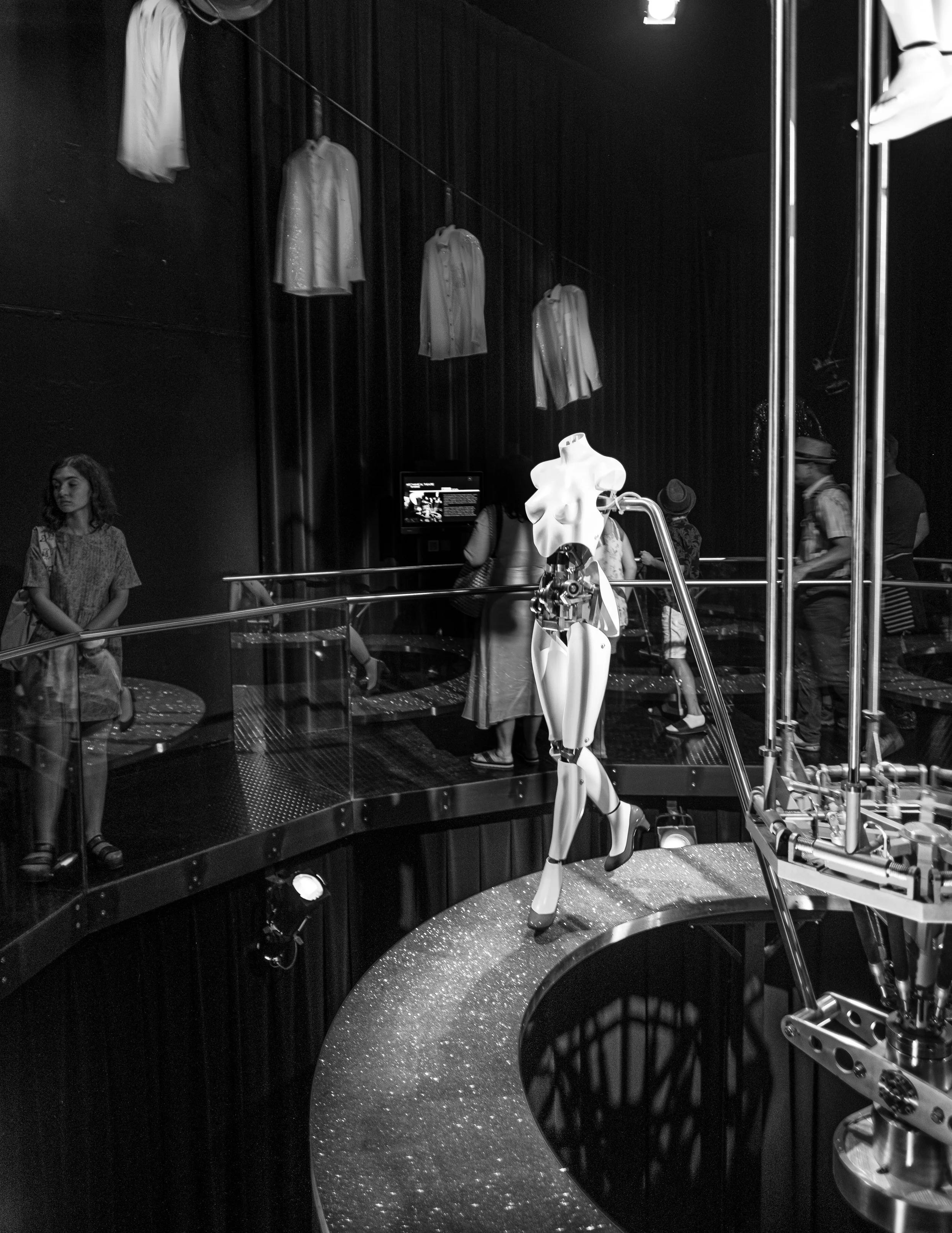 Black and white photo of an art installation in a gallery space. Several white shirts are hanging on a line against a dark curtain backdrop. A mannequin-like sculpture with an abstract, fragmented design is positioned on a circular platform in the fo
