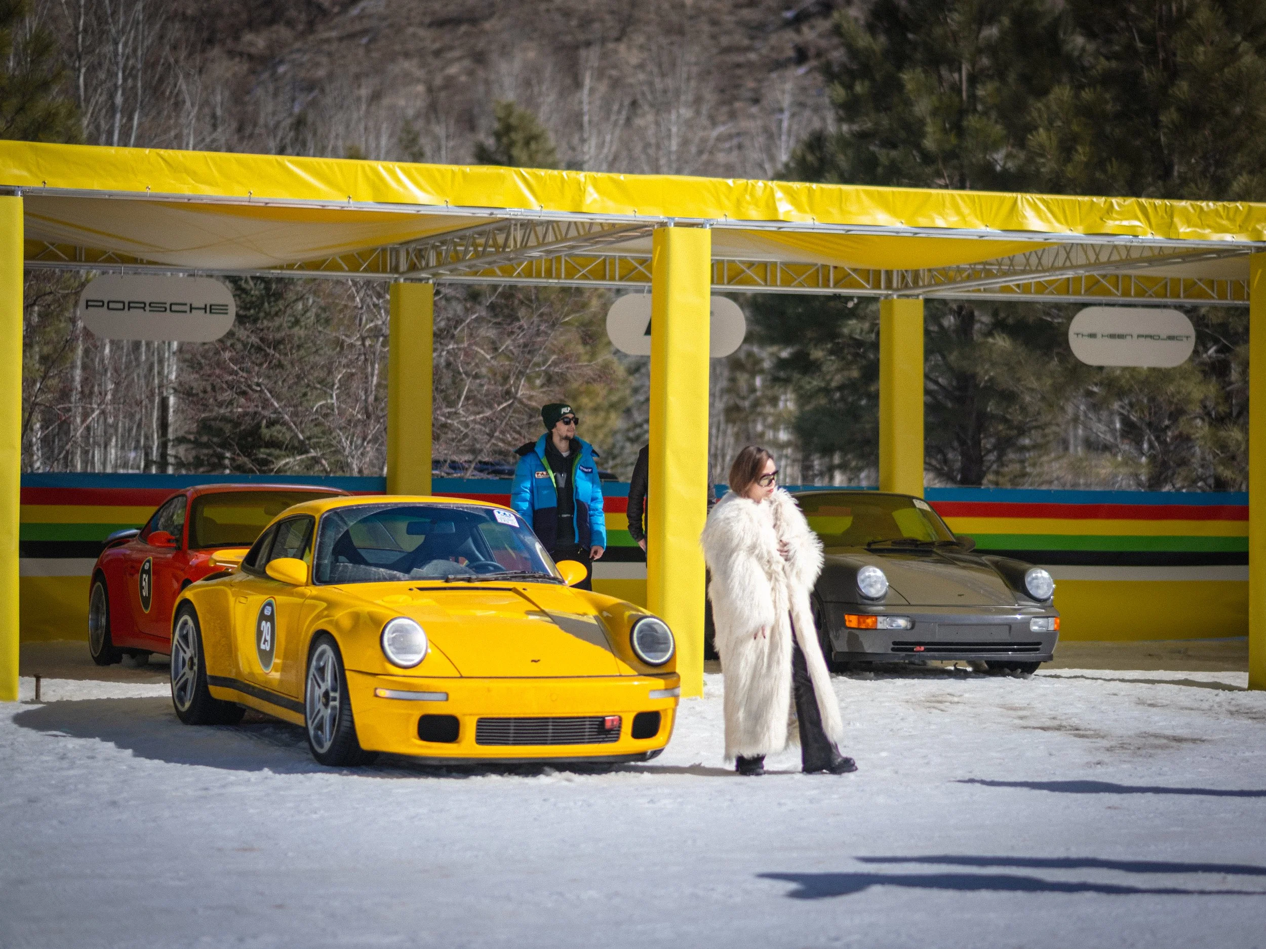 Yellow sports car parked on snow with other colorful sports cars in the background, people walking near the cars, and trees behind a yellow canopy. RUF Porsche, Fat Ice Race, Aspen Colorado