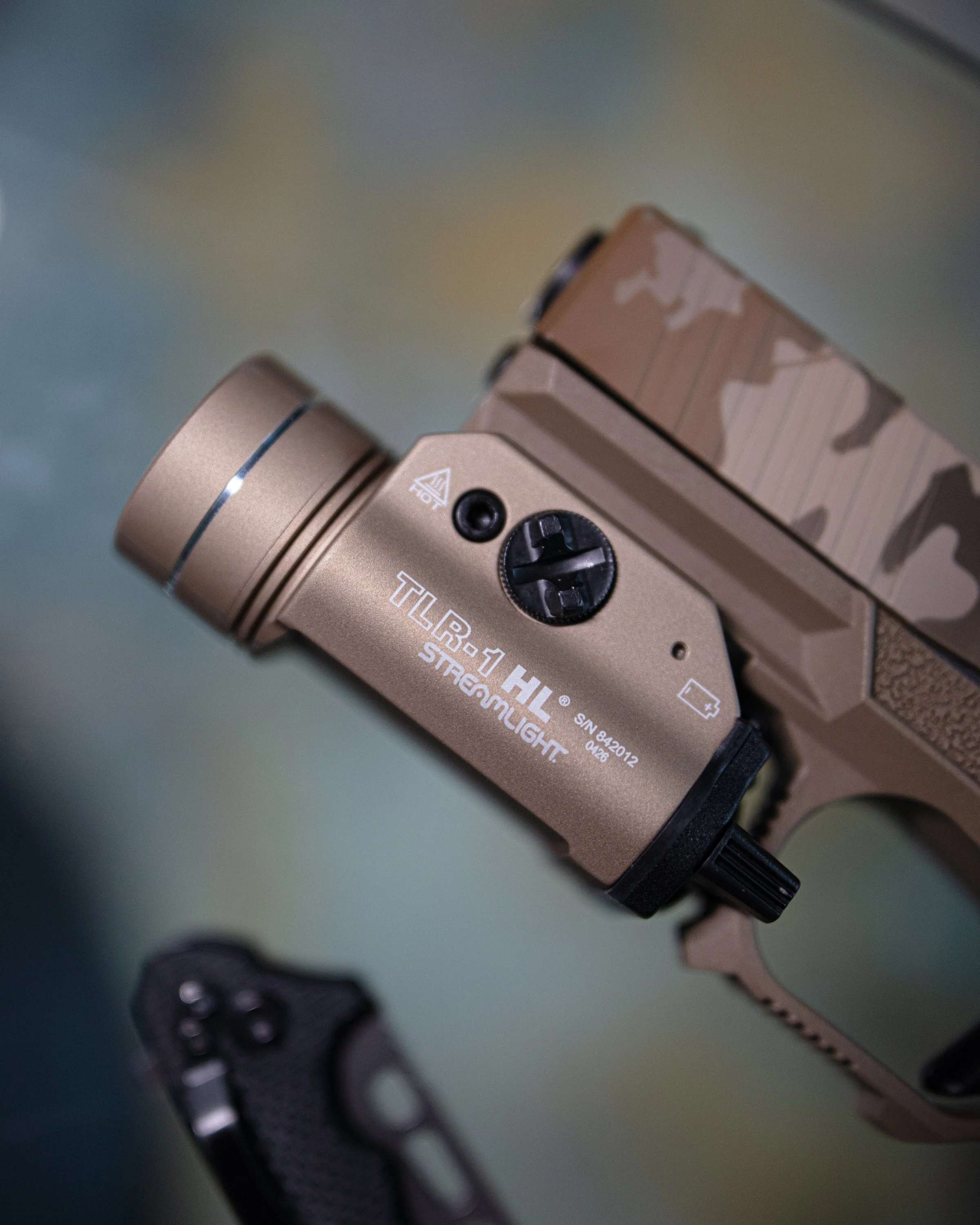 Close-up of a tactical flashlight attached to a camouflage-patterned firearm, with the flashlight's markings and part of the firearm's grip visible.