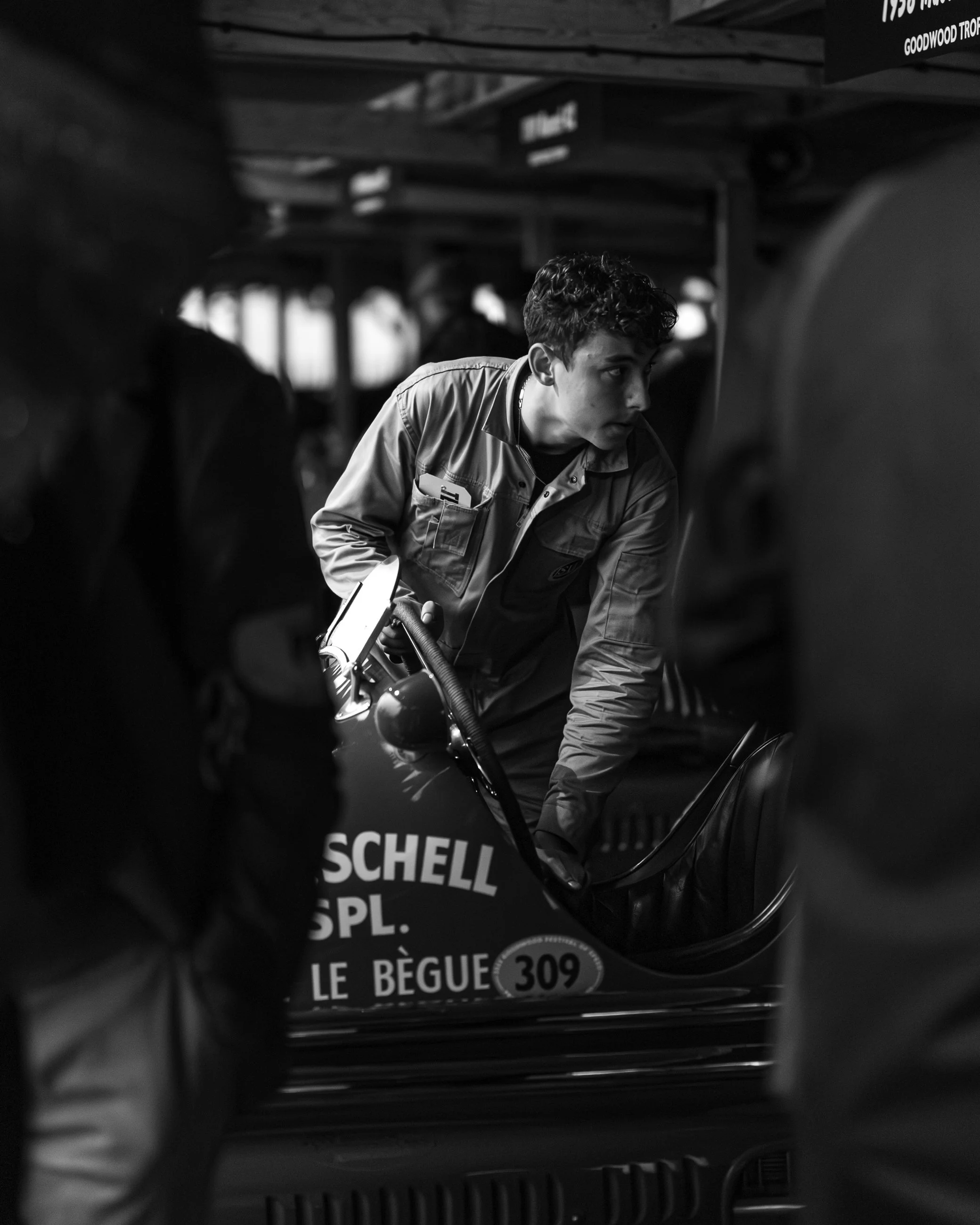 A person with curly hair wearing a jacket looking to the side, inside a vehicle with various signs and labels, including 'Schell' and 'LE BEGUE 309'.