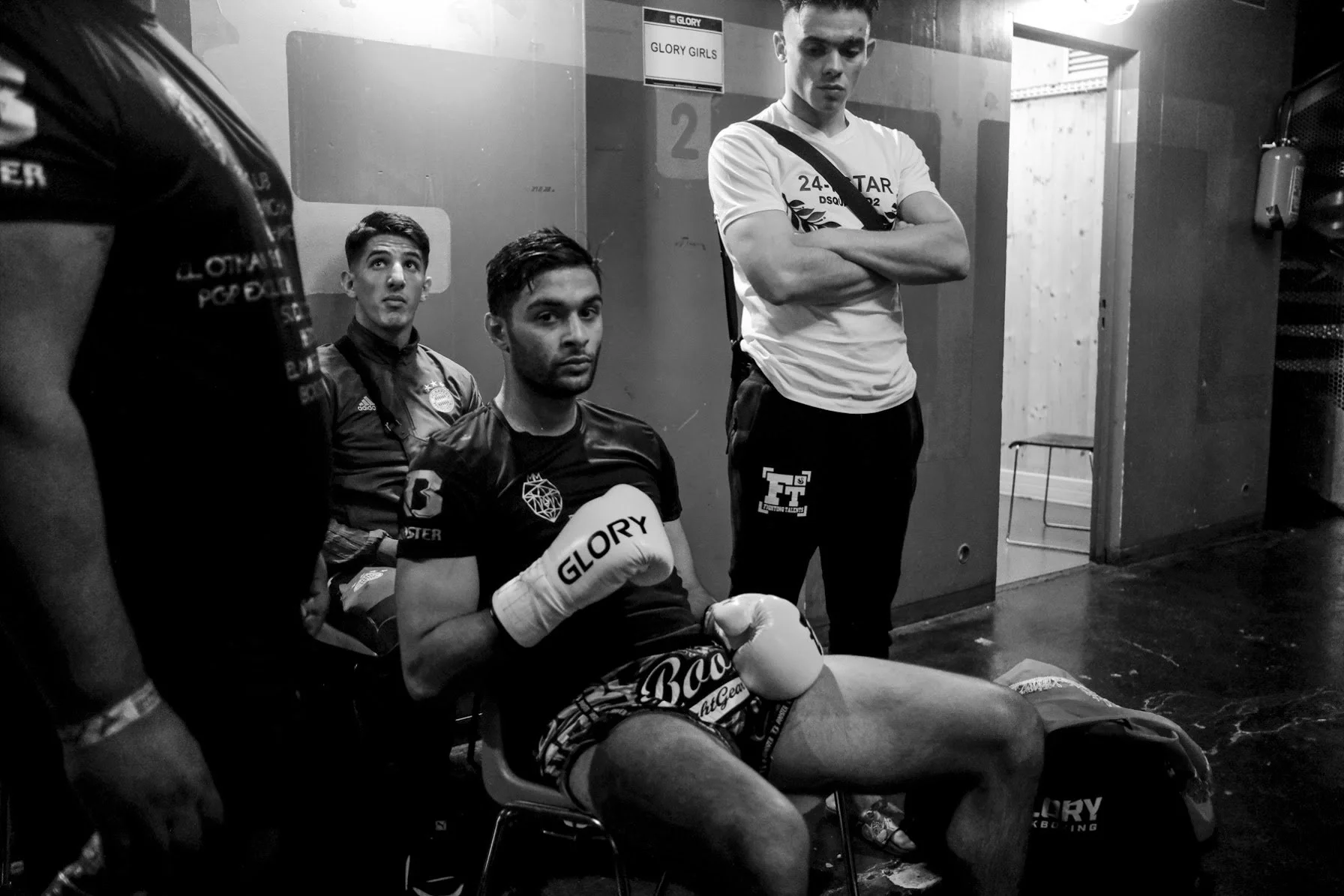 Four young men in boxing attire, two seated and two standing, in a locker room with a sign that reads 'GLORY GIRLS'. The seated man is wearing boxing gloves and shorts, and the standing man has his arms crossed. The others are partially visible.