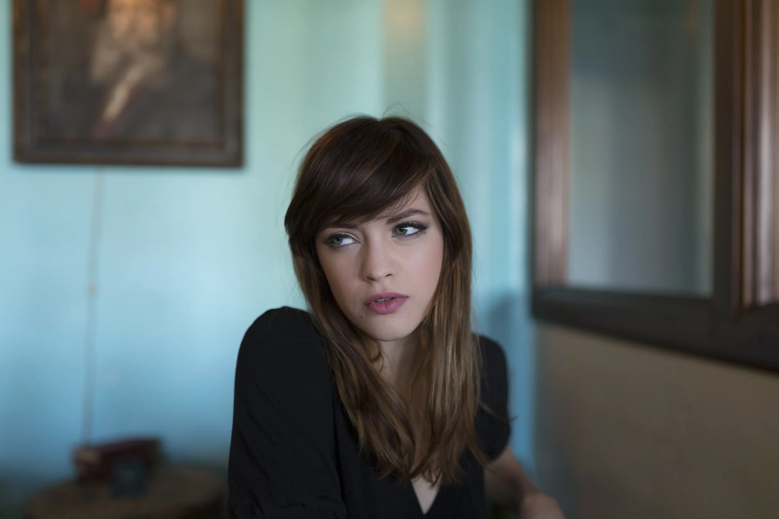 A woman with brown hair looking slightly to her left, sitting in a room with teal walls and framed pictures.