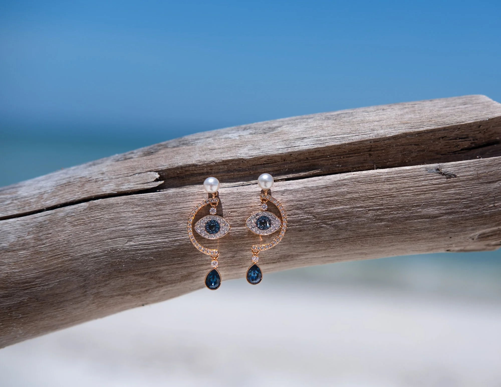 Pearl and black gemstone drop earrings hanging on a weathered wooden surface with a blurred blue sky in the background. Swarovski.