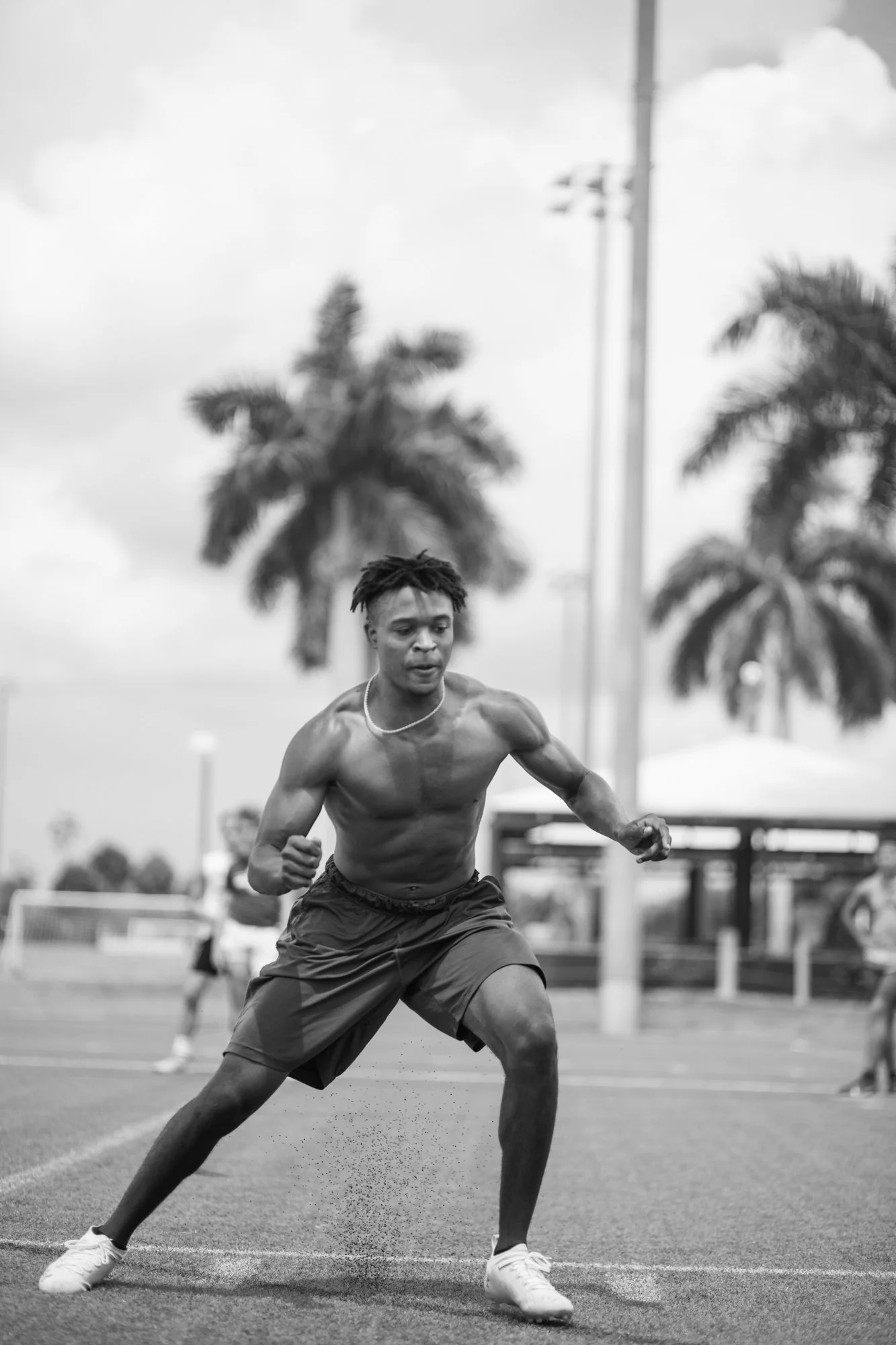 A young male athlete in shorts and white sneakers is performing a sprint start on an outdoor track with palm trees and cloudy sky in the background.