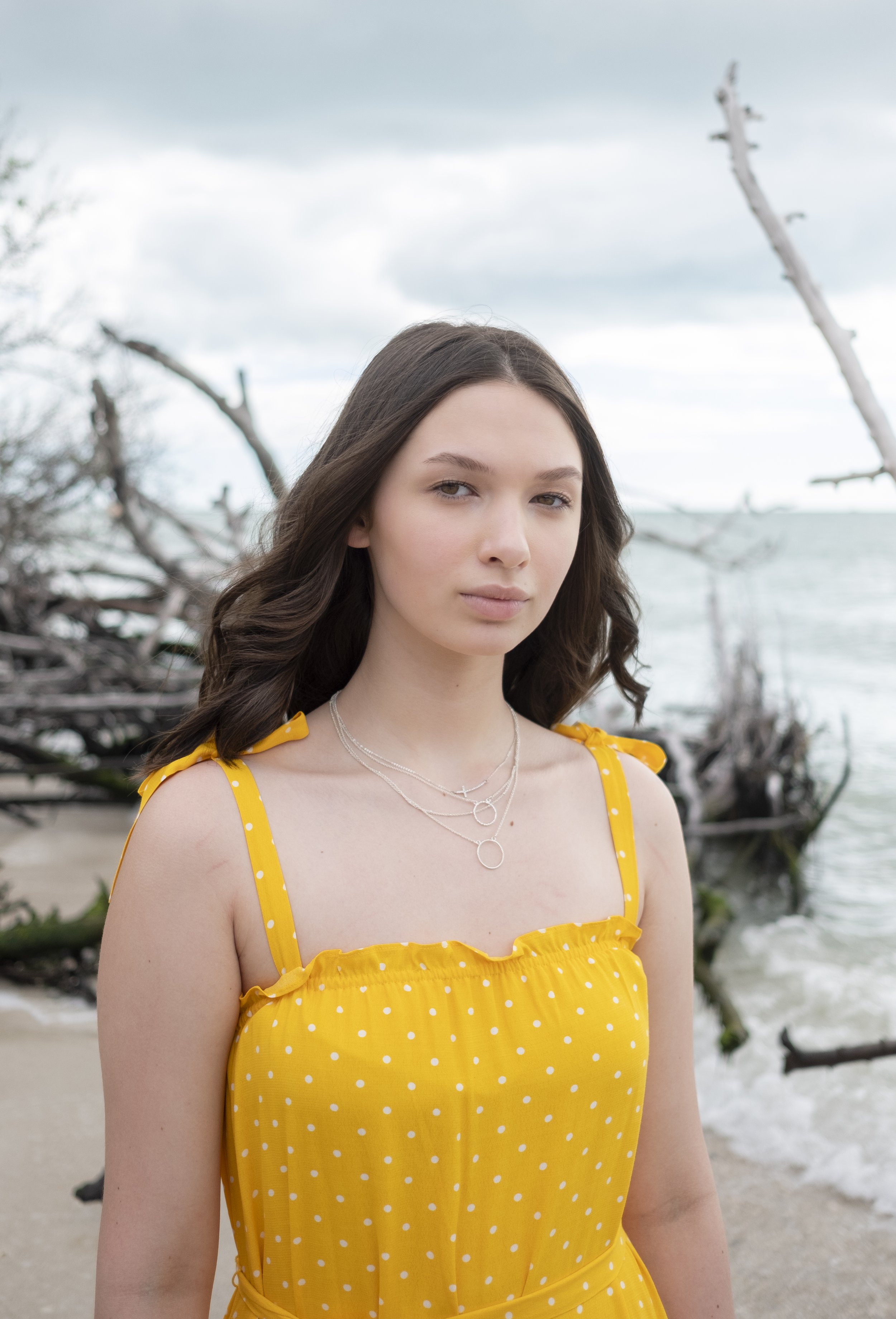 A young woman with long brown hair in a yellow polka dot dress stands on a beach with driftwood and cloudy sky in the background.