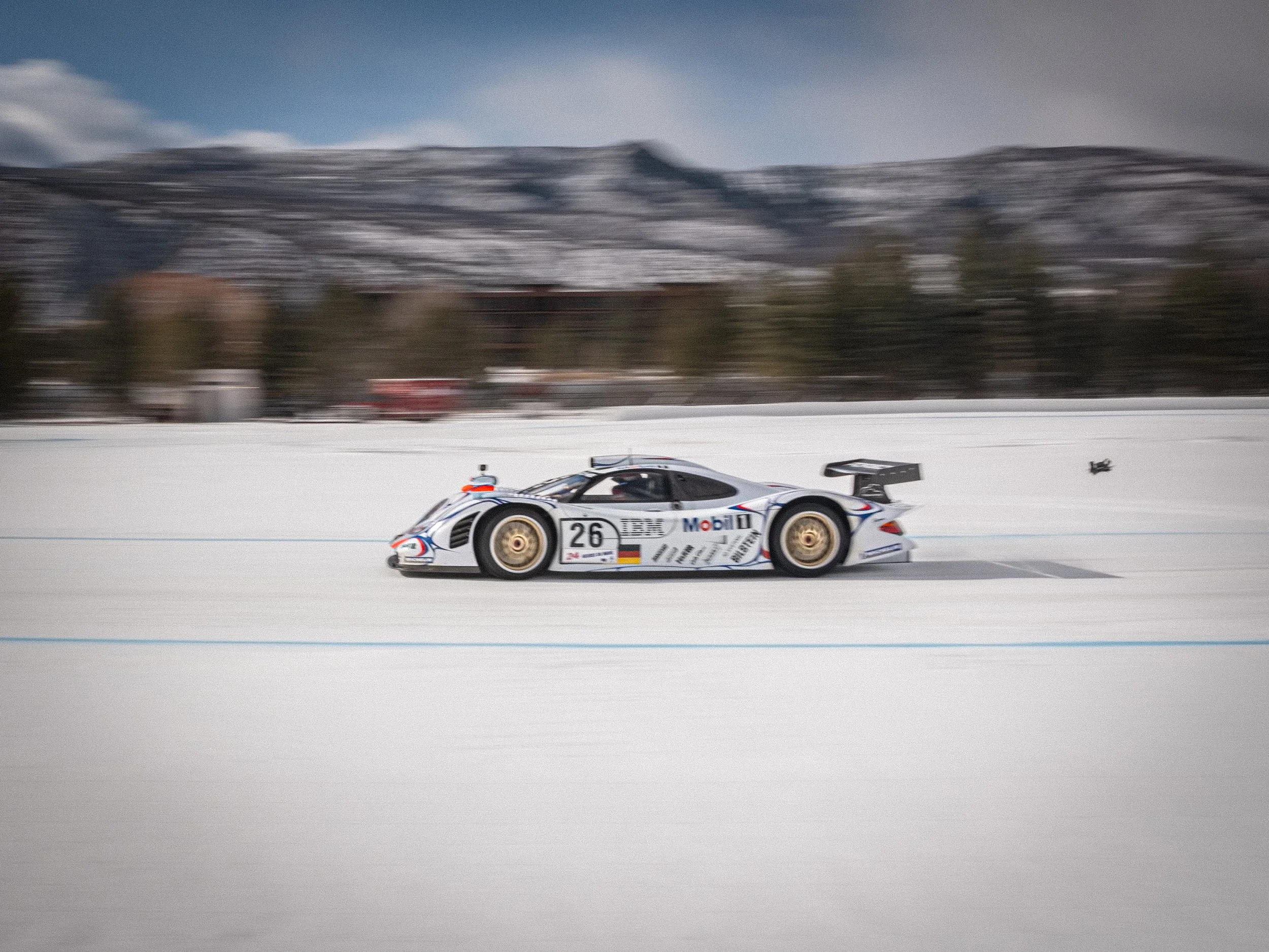 Race car speeding on snow-covered track with mountains in the background. A race car moving on a snowy track with trees and buildings in the background. Porsche LeMans race car Fat Ice Race