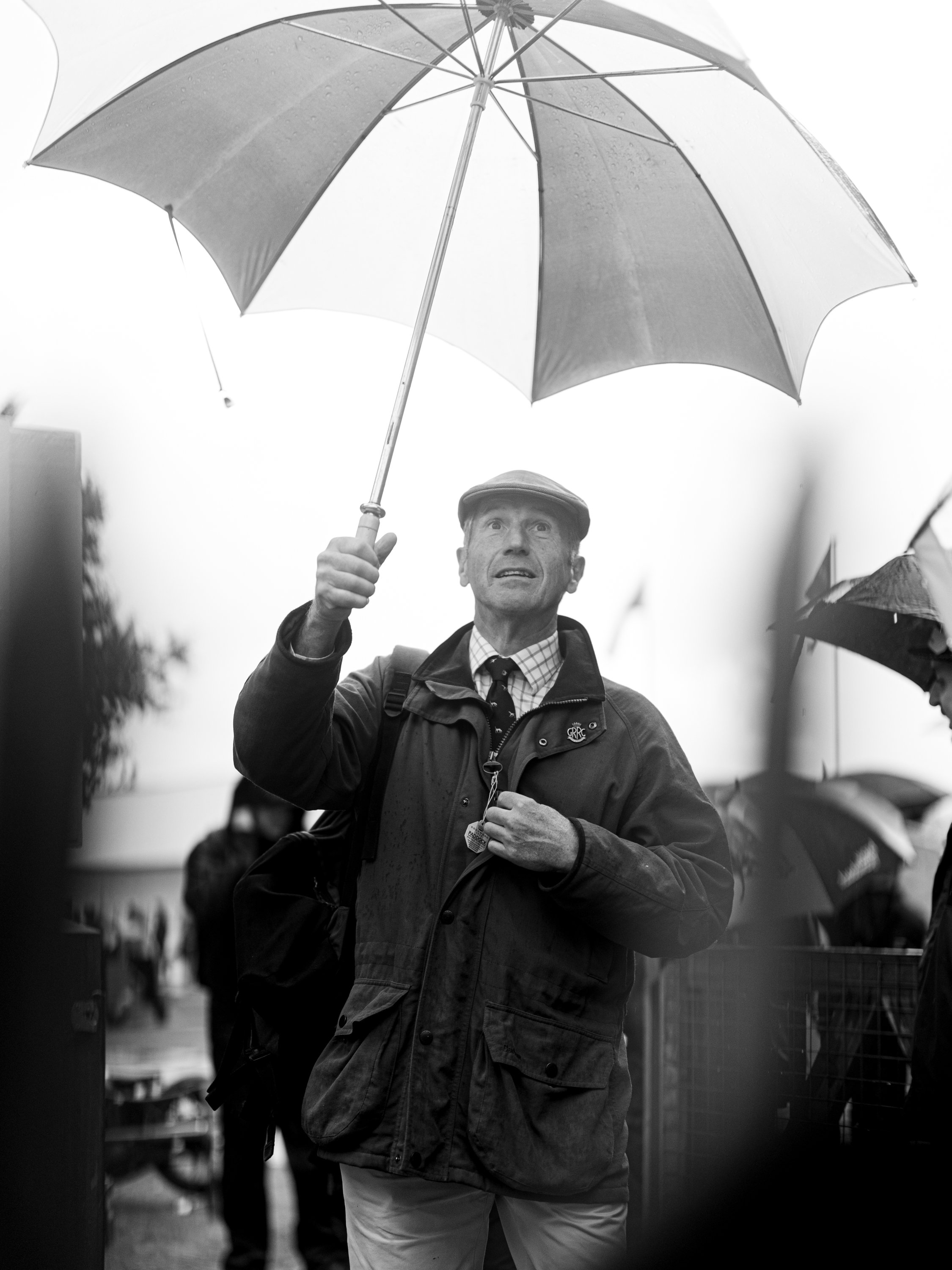 A man holding an umbrella in a crowd, wearing a hat, jacket, and backpack, with other umbrellas nearby.