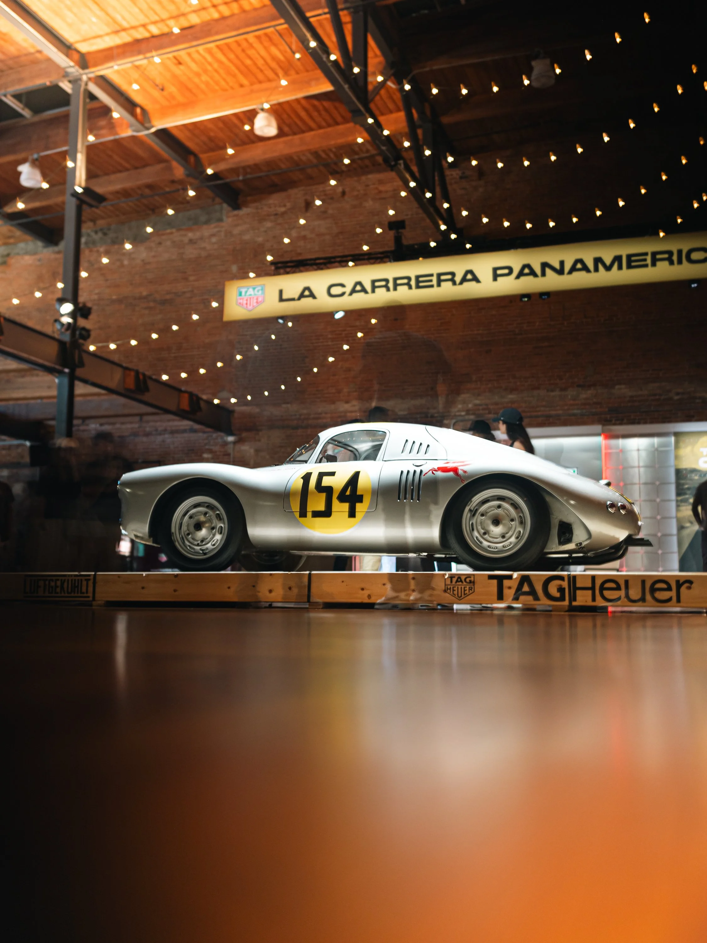 A vintage race car with the number 154 displayed on its side, placed on a wooden platform with TAG Heuer branding in an indoor setting with hanging string lights and a brick wall background.