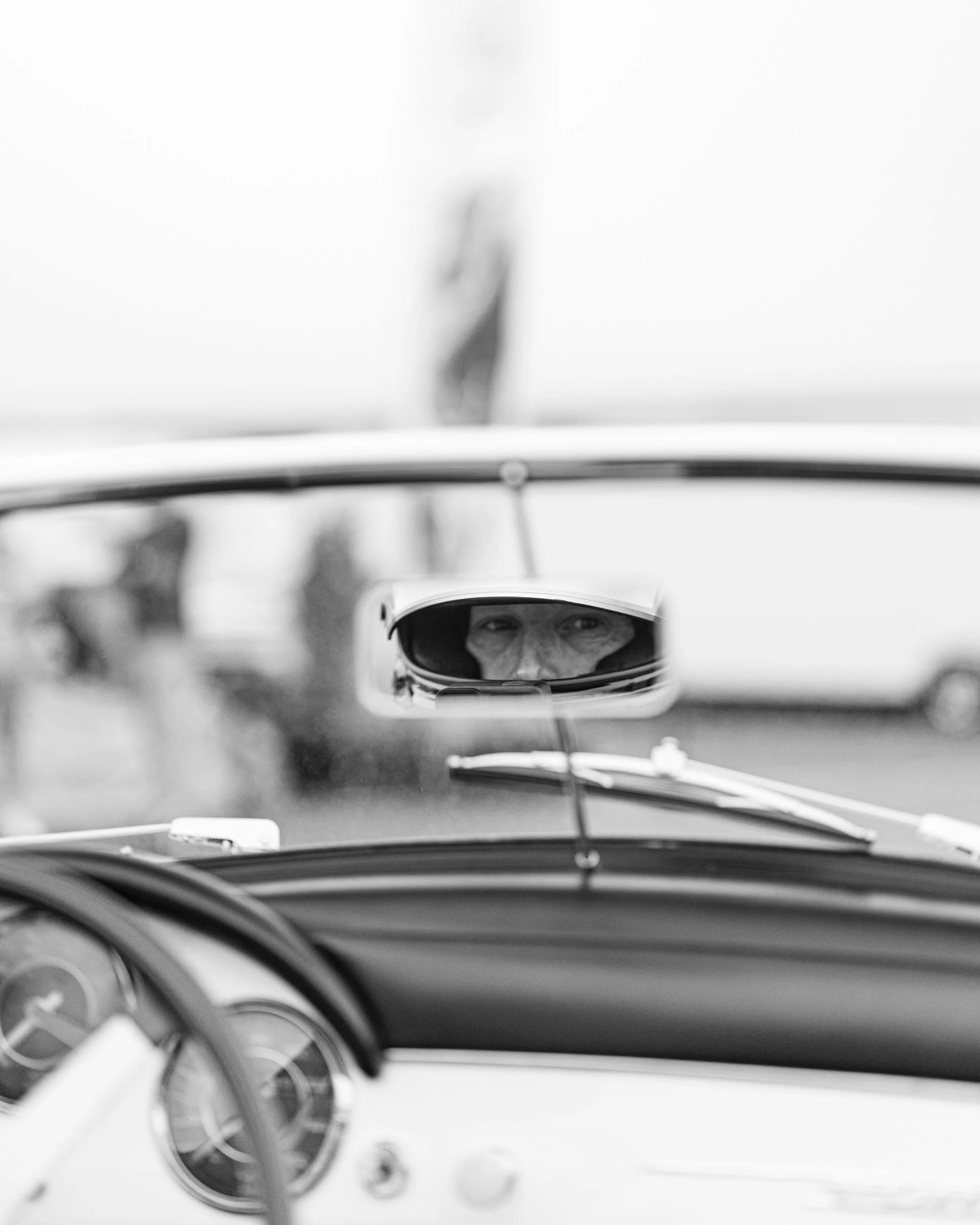 Rearview mirror showing a person's face wearing a helmet inside a race car. The dashboard and steering wheel are visible in the foreground, with blurred gauges and controls.