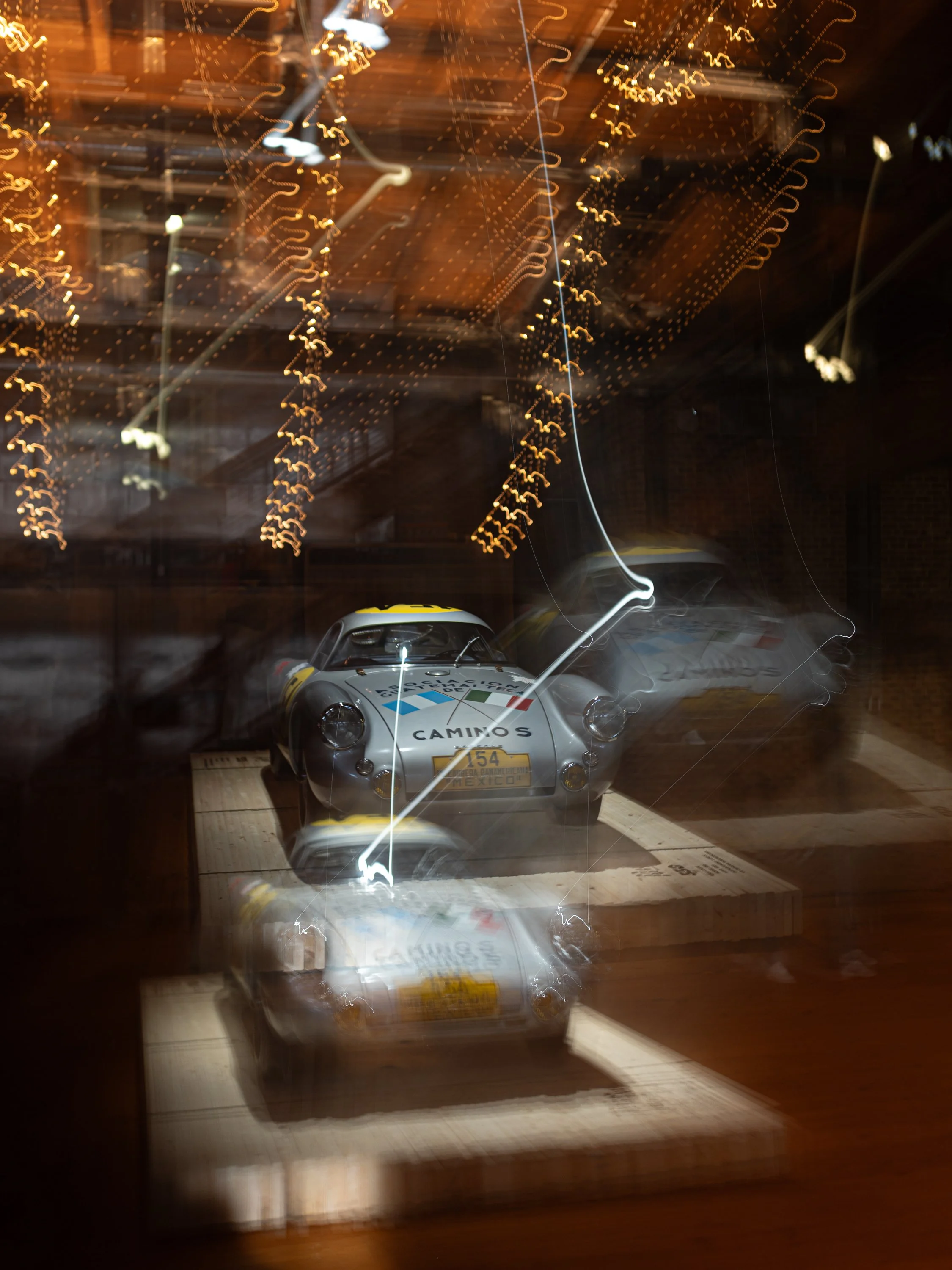 A silver racing car with logos and a yellow license plate, on display on a wooden platform with reflective glass, surrounded by blurred lights and streaks from camera movement.