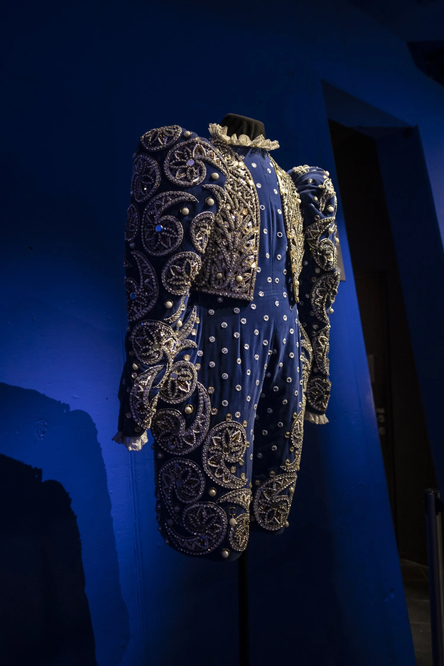 A mannequin dressed in an ornate, embroidered suit with intricate beadwork and pearl accents, displayed against a blue wall with dramatic lighting. Swarovski.