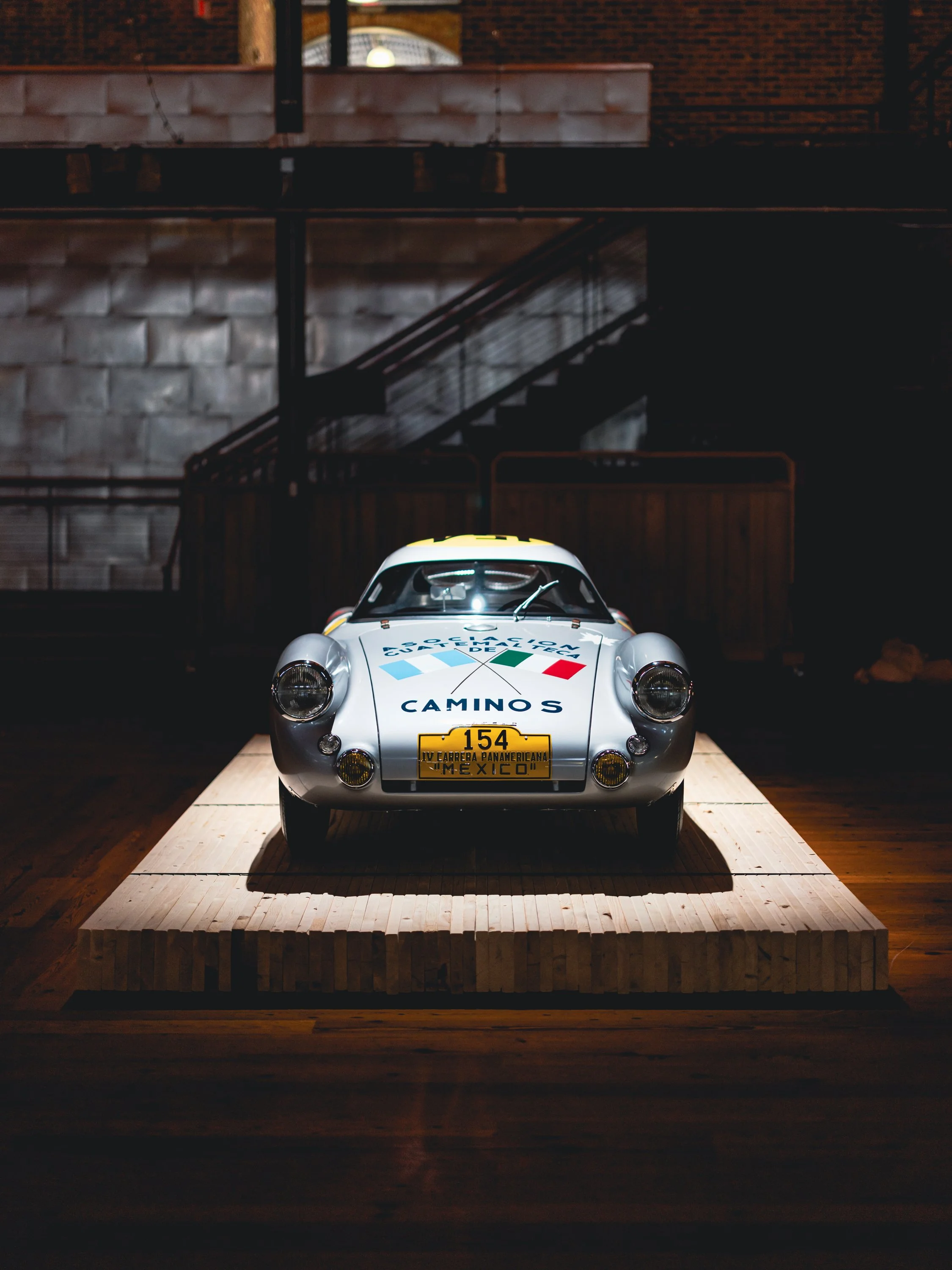 A vintage race car displayed on a wooden platform inside a dimly lit building, with brick and wooden interior in the background. luftgekuhlt