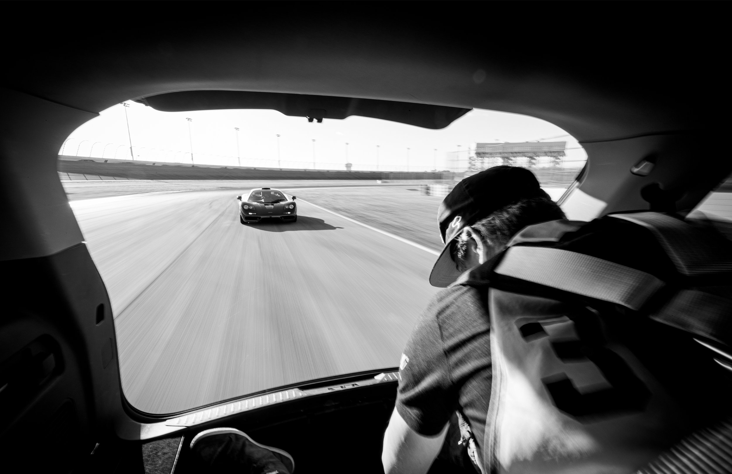 Black-and-white photo of a race car driver and crew member inside a race vehicle, racing on a track with another race car ahead.