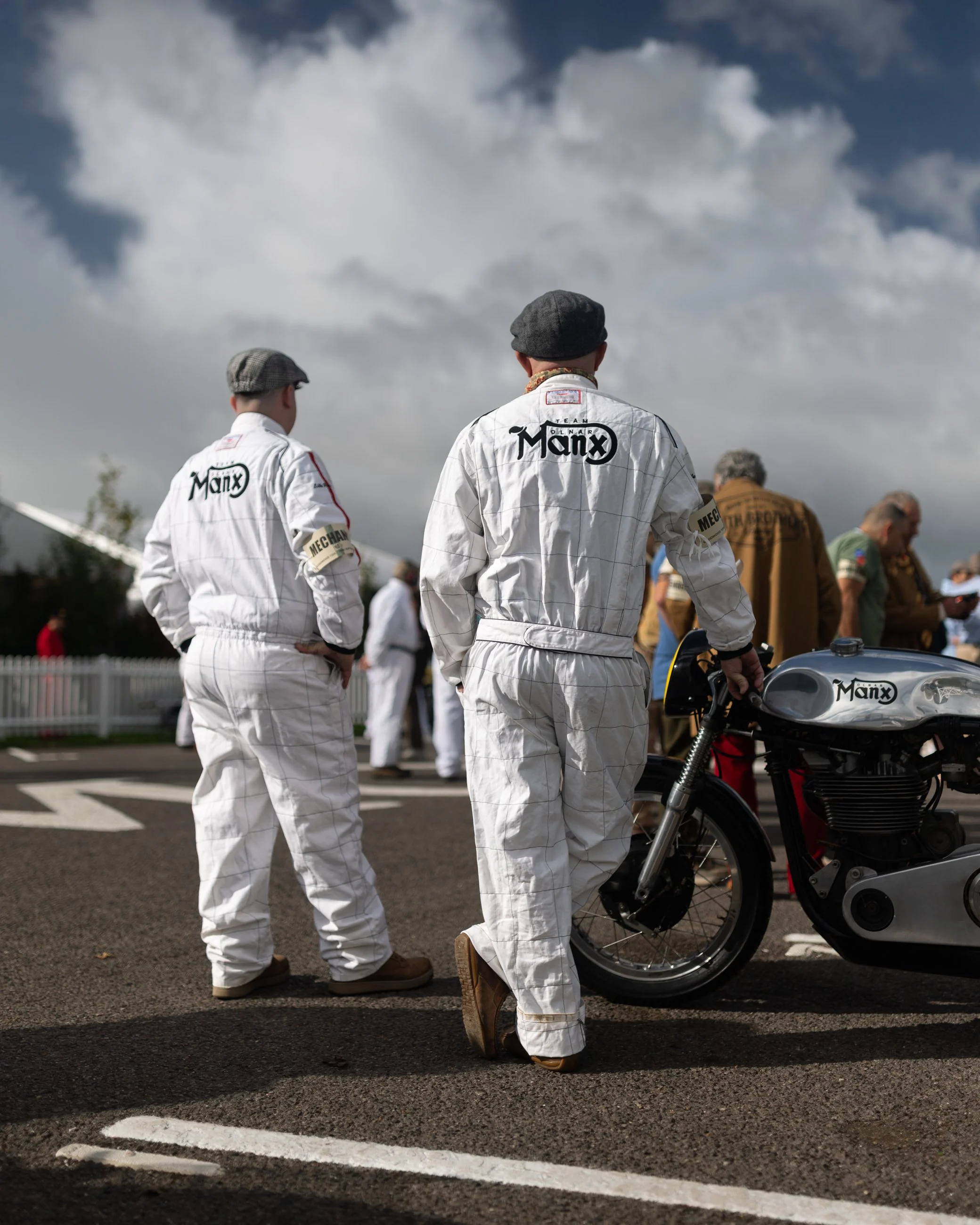 Two men dressed in racing suits and flat caps standing next to a motorcycle in a parking lot, with other people and cloudy sky in the background. Goodwood Revival.