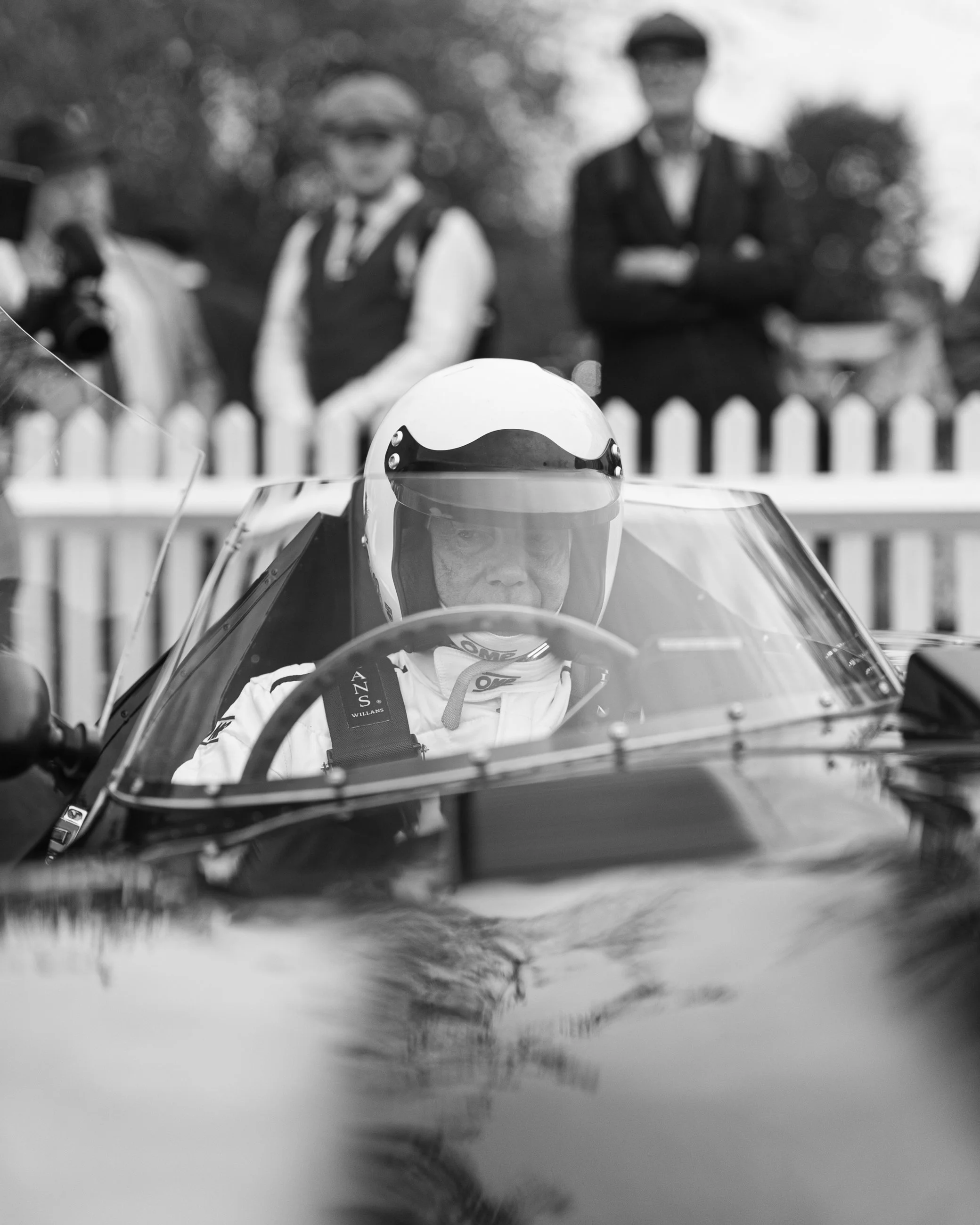 A person wearing a racing helmet and racing suit sitting in a racing car, with a white picket fence and trees in the background.
