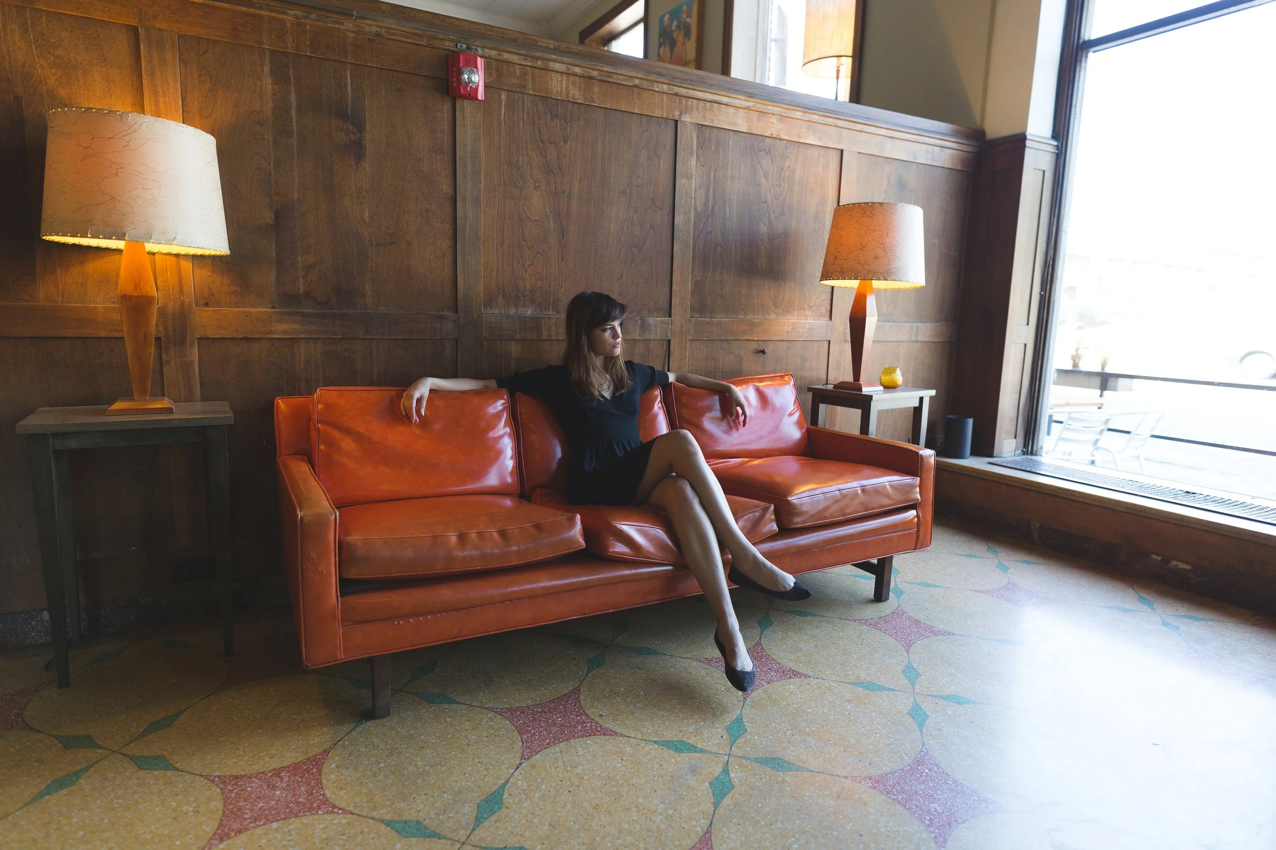 A woman sitting on a burnt orange leather sofa in a room with wooden paneling on the wall and large windows. She appears thoughtful, dressed in a black dress with her legs crossed and arms resting on the sofa. There are two standing lamps with beige 