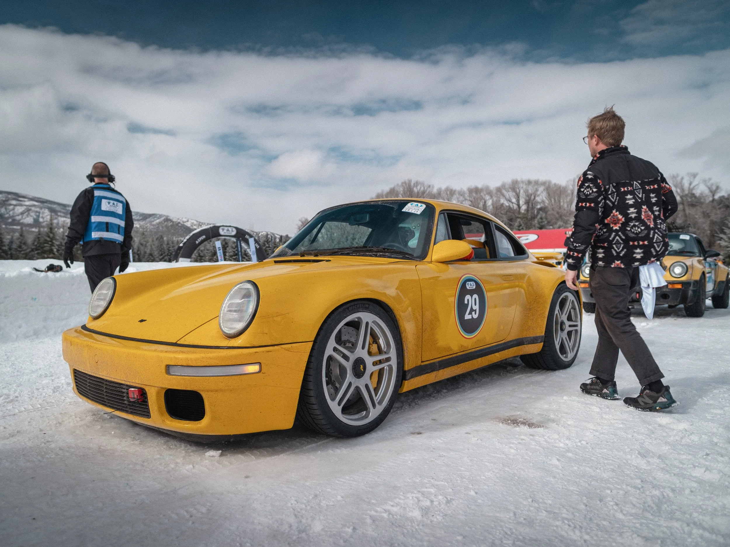 A yellow sports car with the number 29 parked on a snowy landscape, with two people nearby, one wearing a black jacket with a patterned design and the other wearing a blue vest and headphones, during daytime with cloudy sky. RUF Porsche yellowbird.