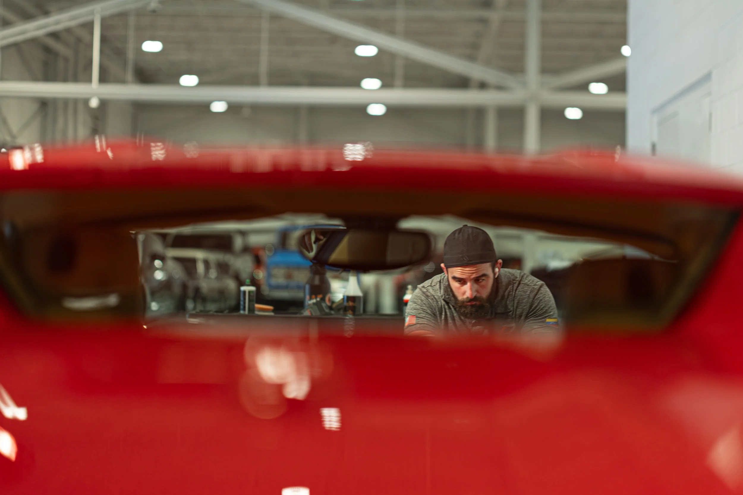 A man with a beard and hat working on a red car in a garage or workshop.