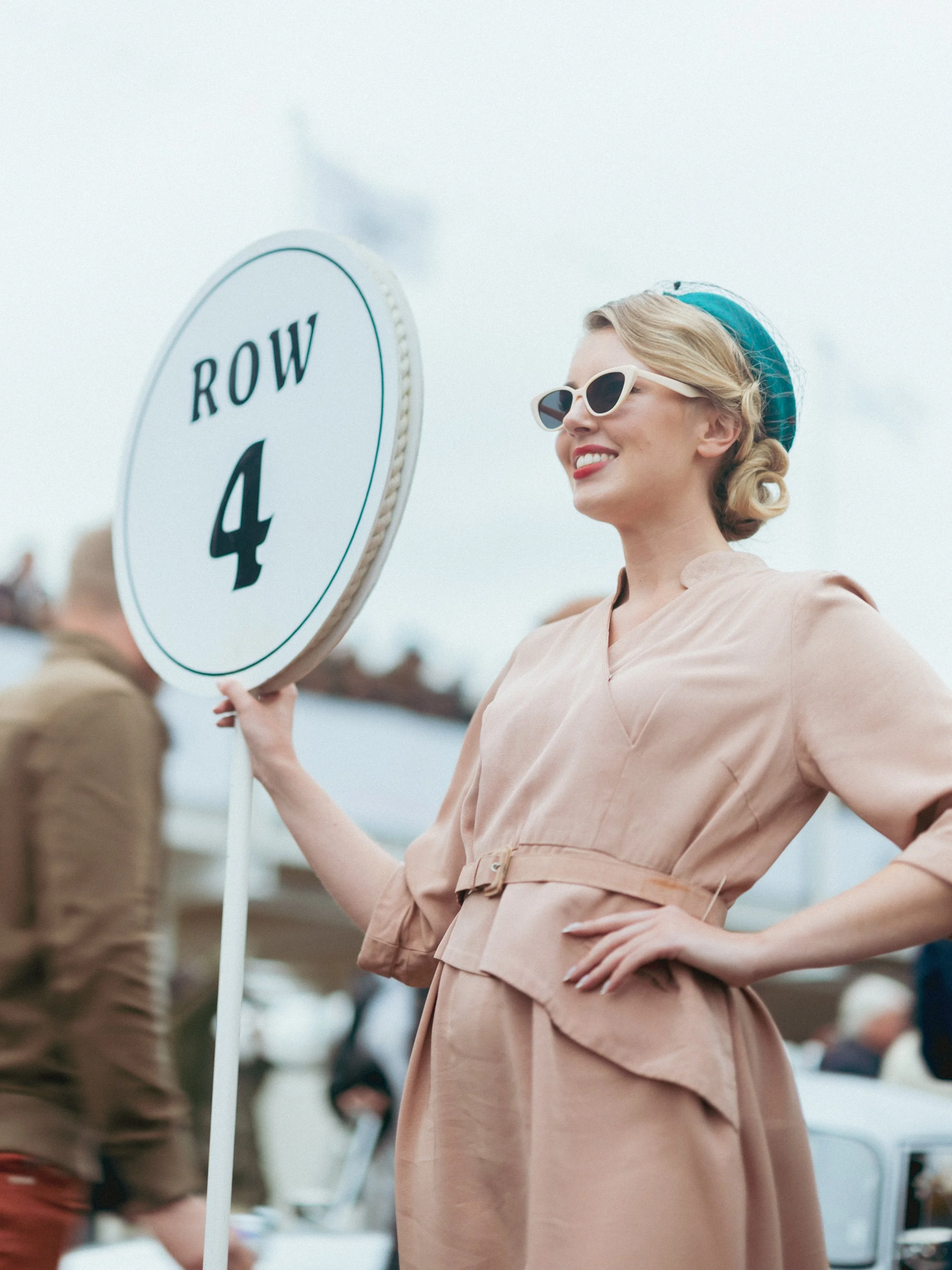 A woman with blonde hair wearing vintage-style sunglasses, a teal headscarf, and a beige dress, holding a large white sign that reads 'ROW 4' at an outdoor event. Goodwood Revival.