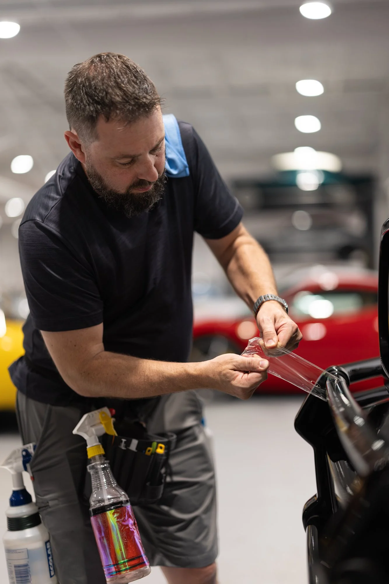 A man in a black shirt and gray shorts is applying a protective film to a black car in a well-lit garage or showroom, with various cleaning and detailing supplies attached to his belt.