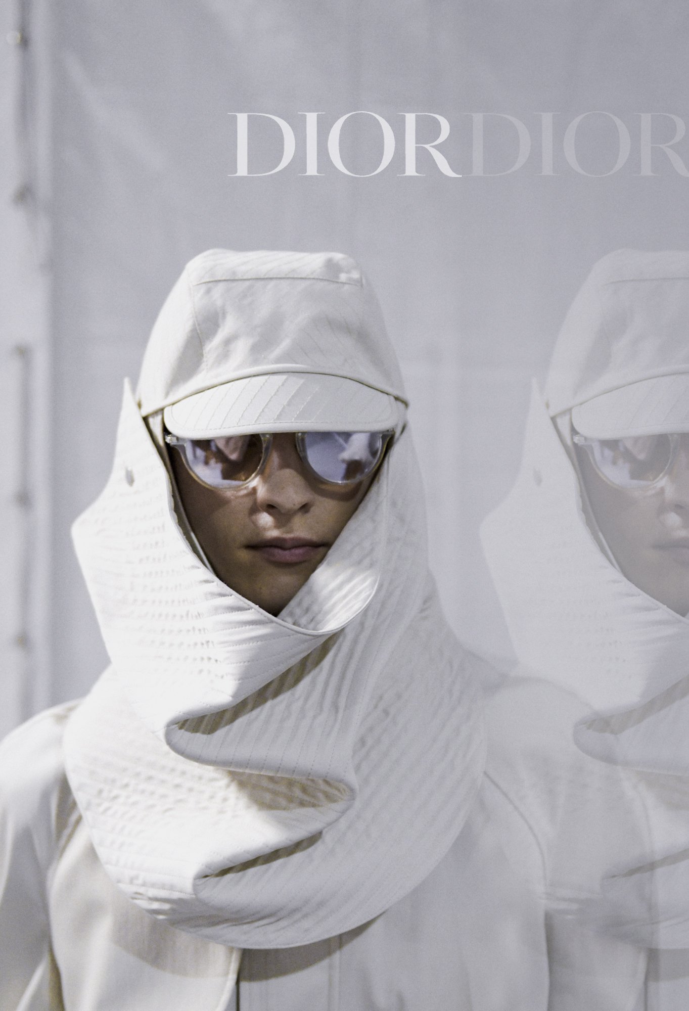 A person dressed in white with a hood and glasses, standing in front of a white background and looking at the camera.