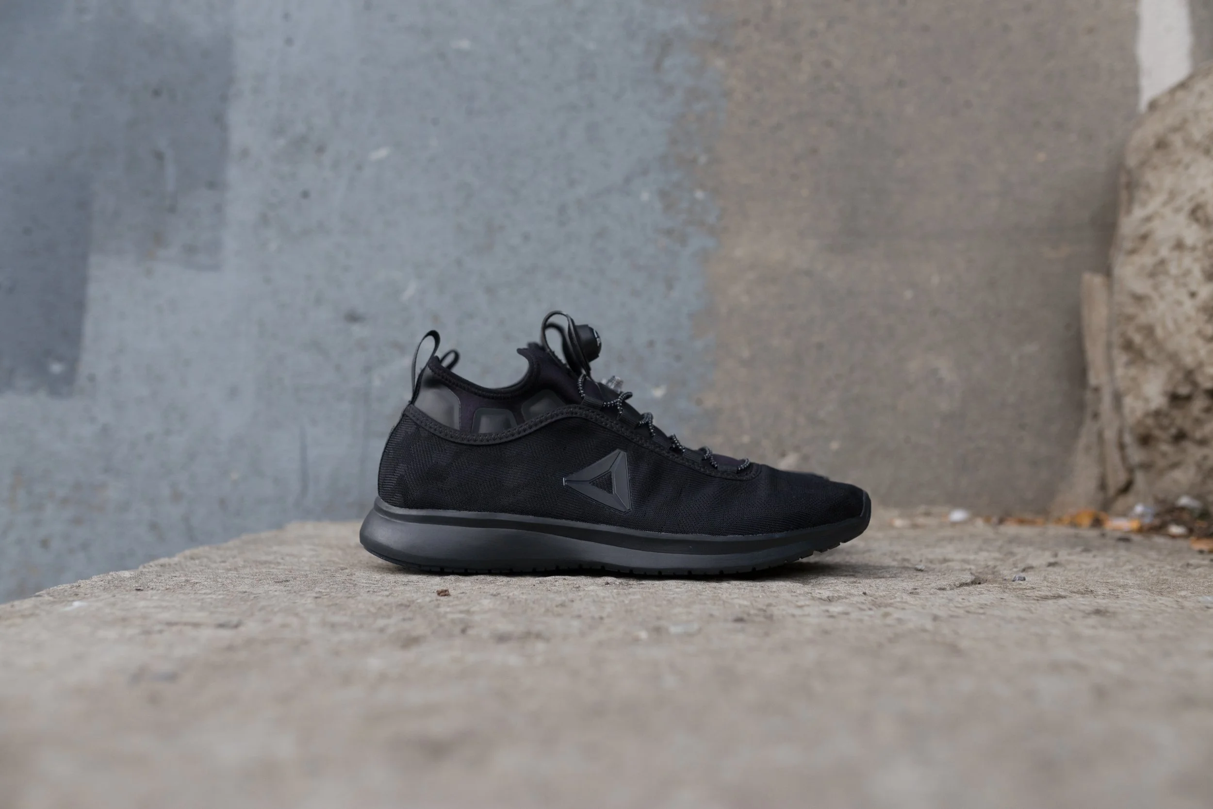 Black athletic shoe with a gray sole on a concrete ground.