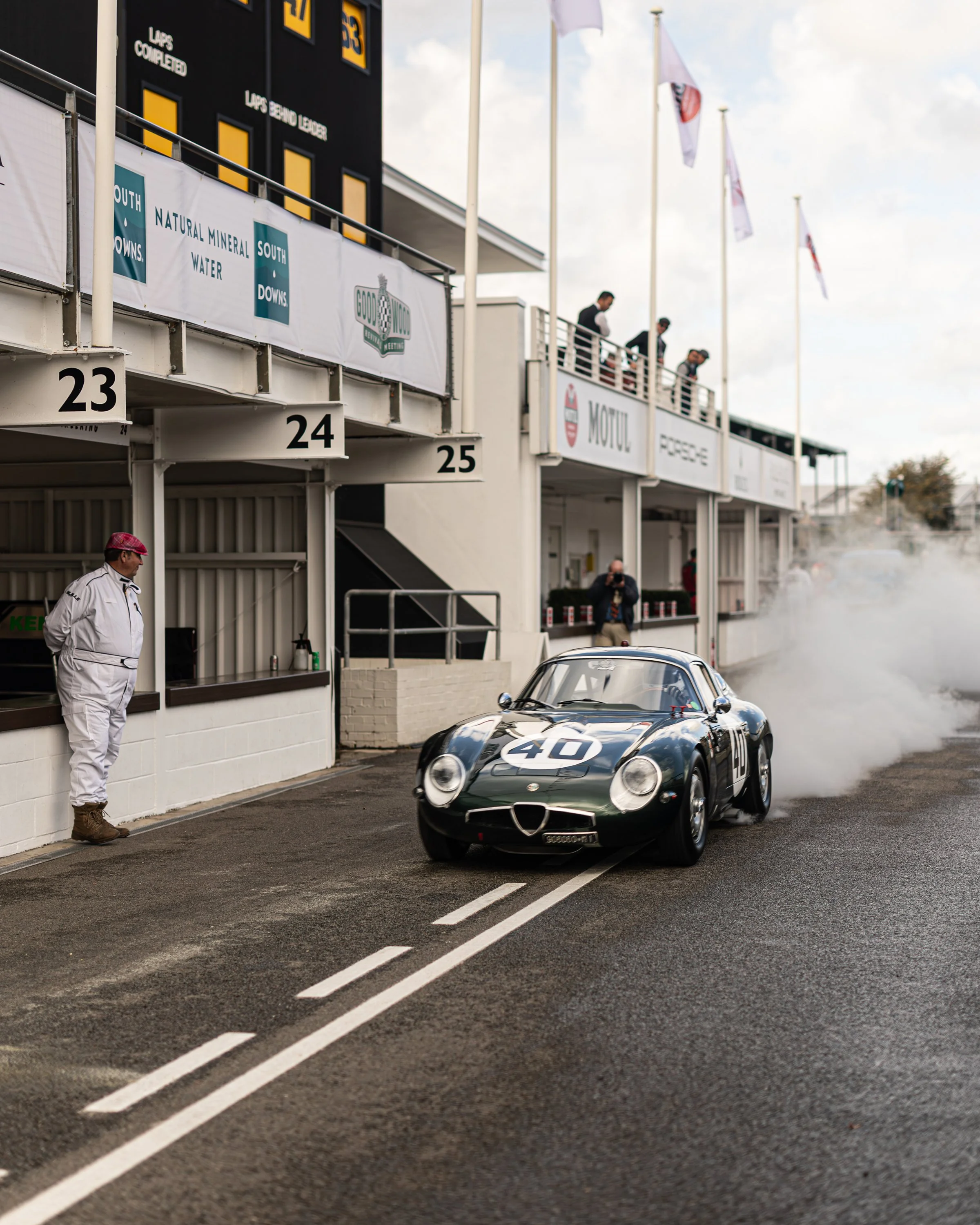 A vintage racing car with the number 40 on the hood, on a racetrack with a cloud of tire smoke behind it, racing past the pit lane with staff and officials observing.