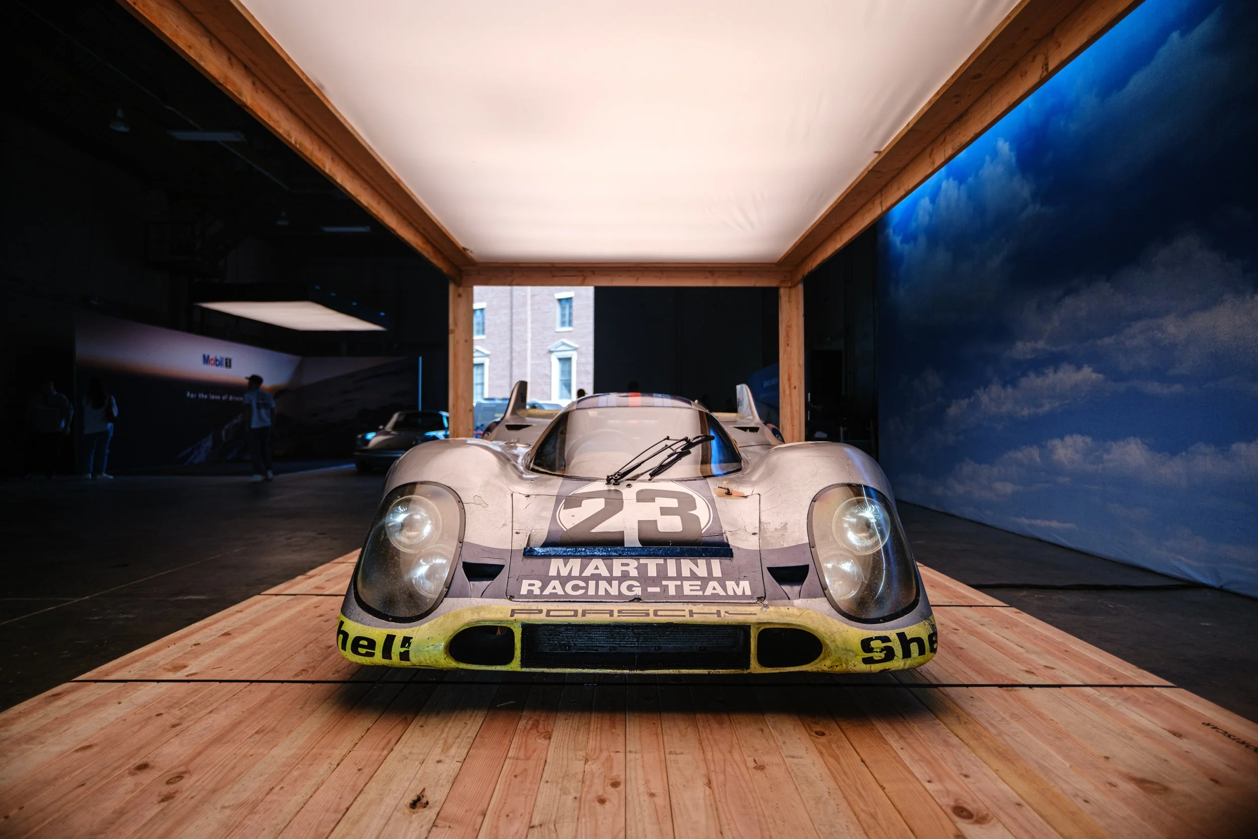 Vintage Porsche race car with Martini Racing Team livery displayed indoors, surrounded by wooden flooring and dark walls, with a large cloud mural on the right wall and other race cars in the background. luftgekuhlt