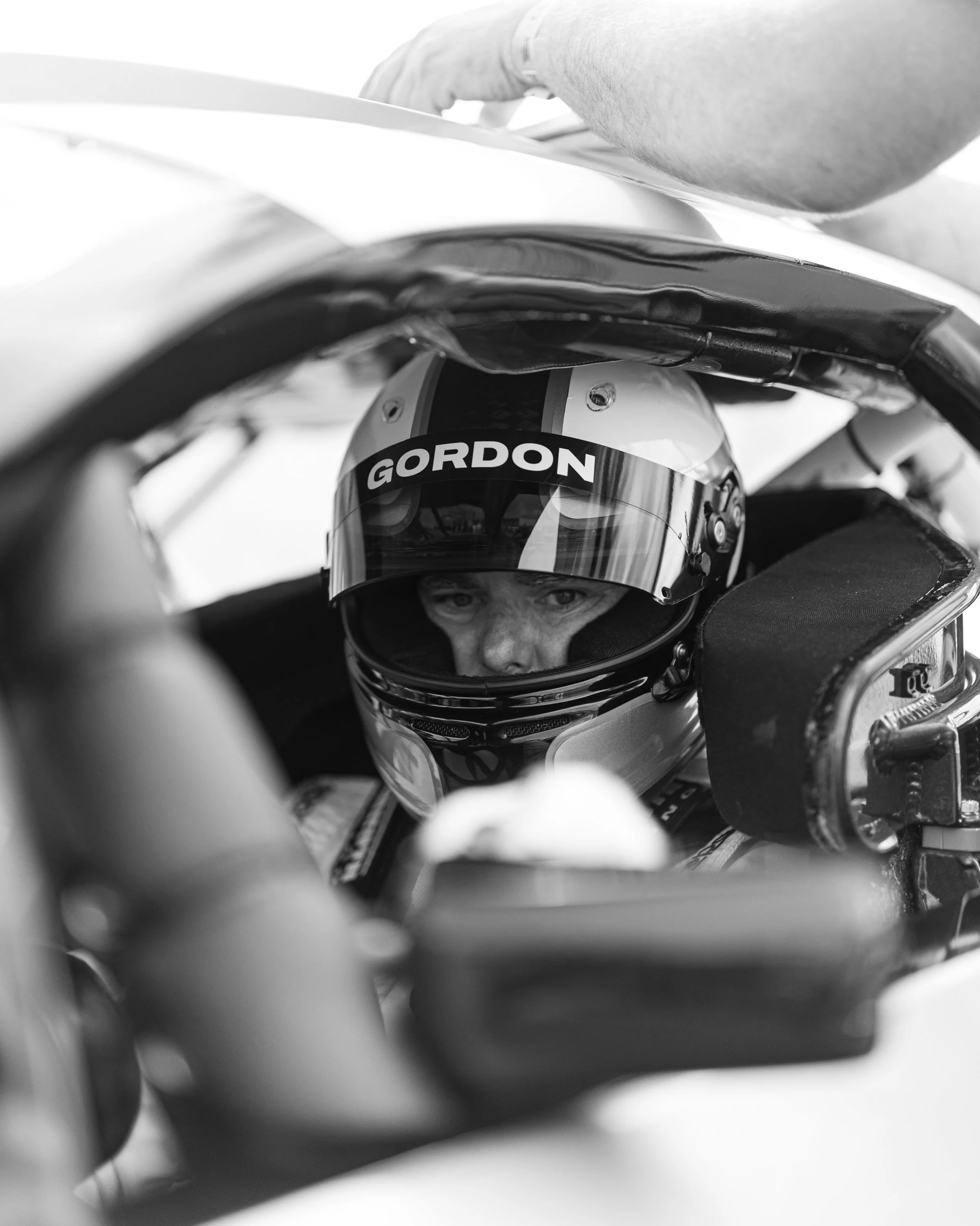 A person wearing a racing helmet with the word 'GORDON' on it, viewed from within a vehicle or a confined space, surrounded by various mechanical and steering components. Jeff Gordon.